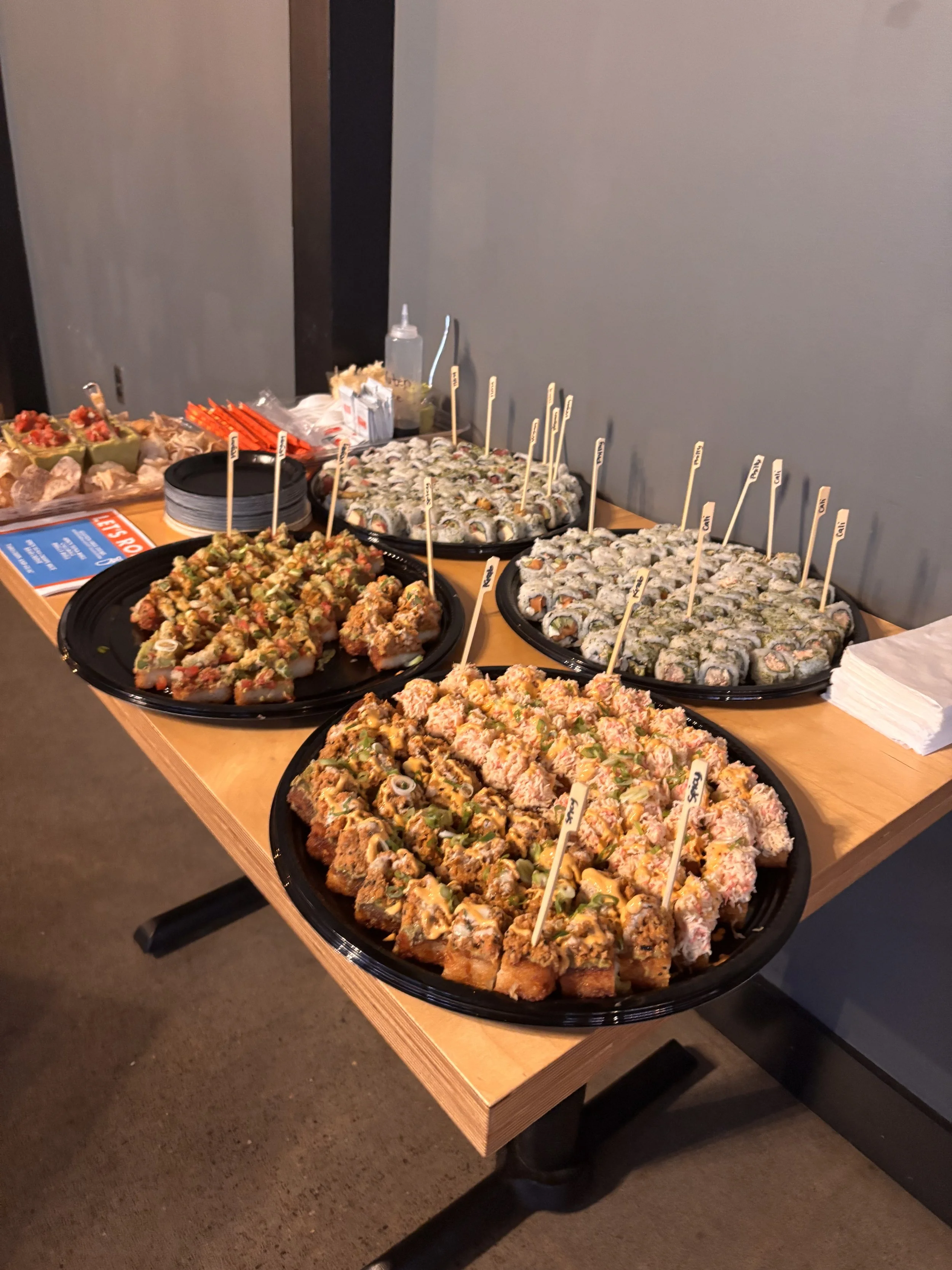 A table with trays of assorted sushi rolls and small appetizers, including labels on picks for different sushi varieties.