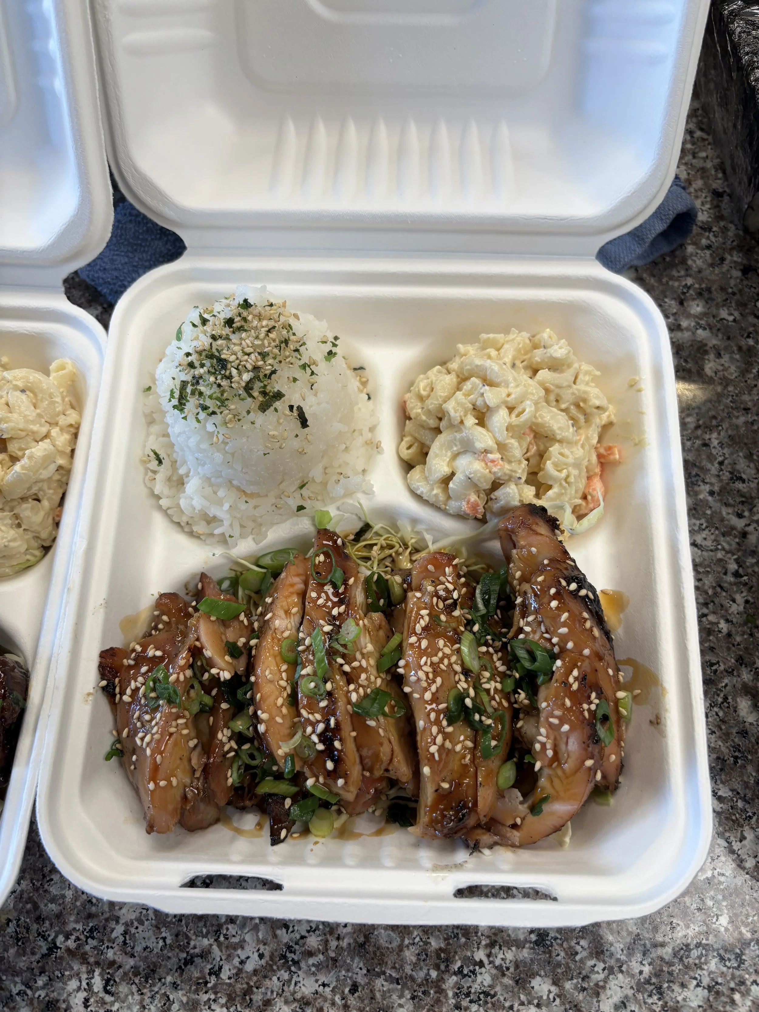 Asian-style chicken with green onions and sesame seeds, served with rice topped with furikake, macaroni salad, and coleslaw in a takeout box.