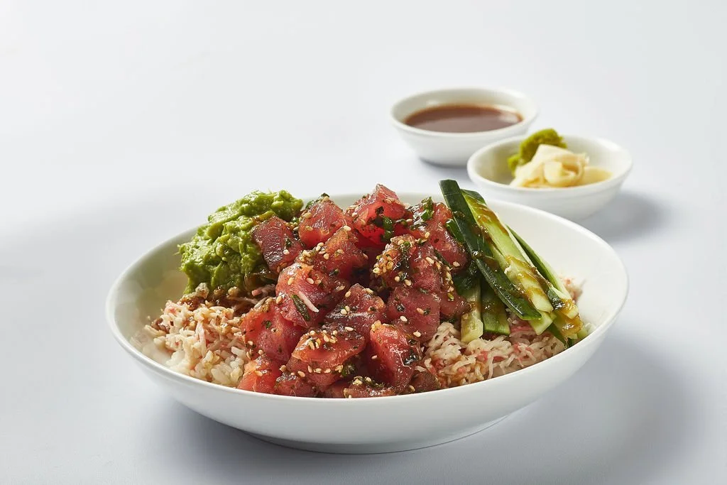 Bowl of poke with diced raw tuna, rice, sliced cucumbers, guacamole, and sesame seeds, with soy sauce, wasabi, and ginger on the side.