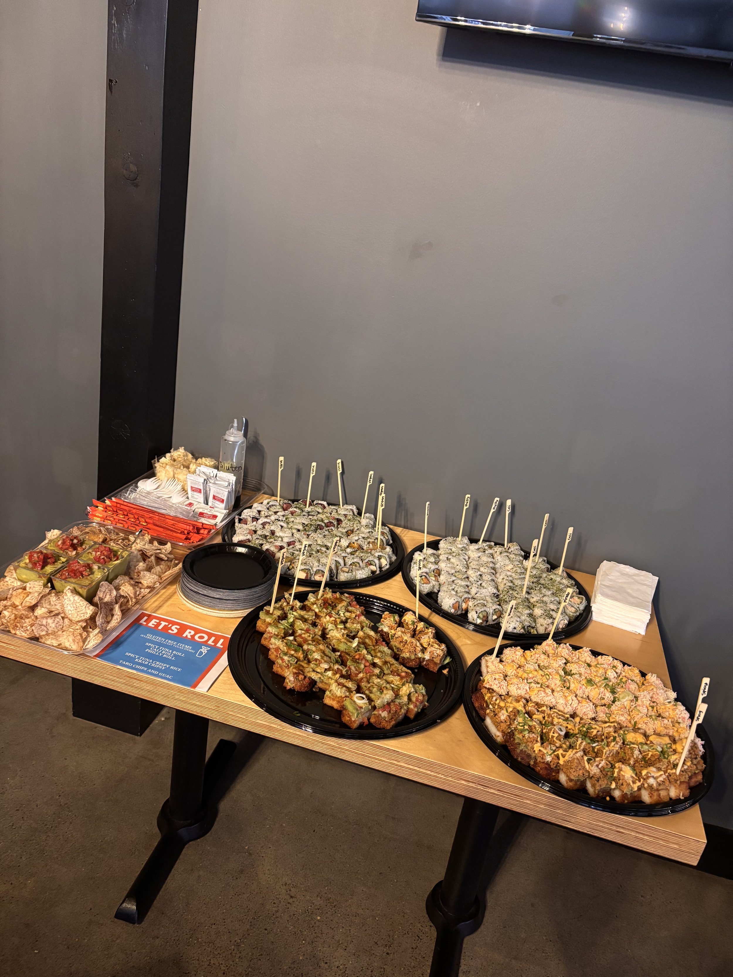 Assorted sushi platters and snacks on a wooden table at an indoor event.
