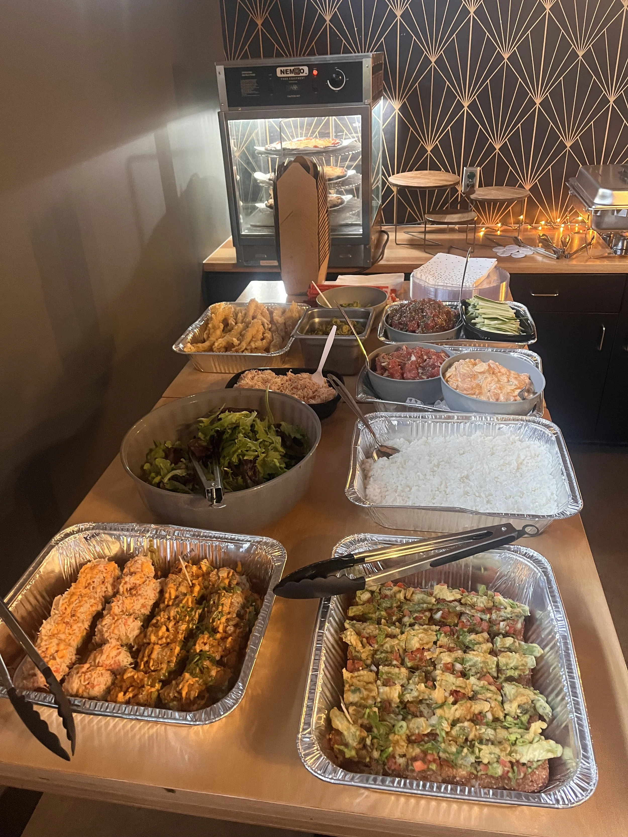 Buffet table with various dishes including salads, sushi rolls, rice, fried chicken, and other appetizers, with an oven in the background.