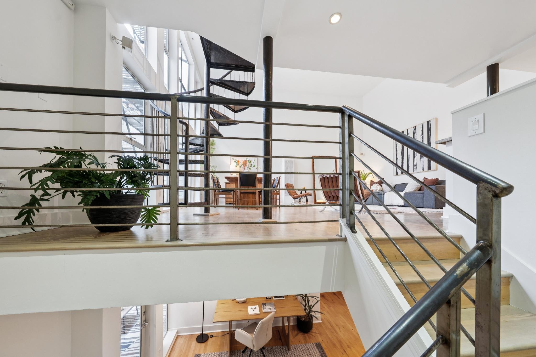 Interior of a modern multi-level home with a metal railing, wooden flooring, potted plants, white walls, and a spiral staircase.