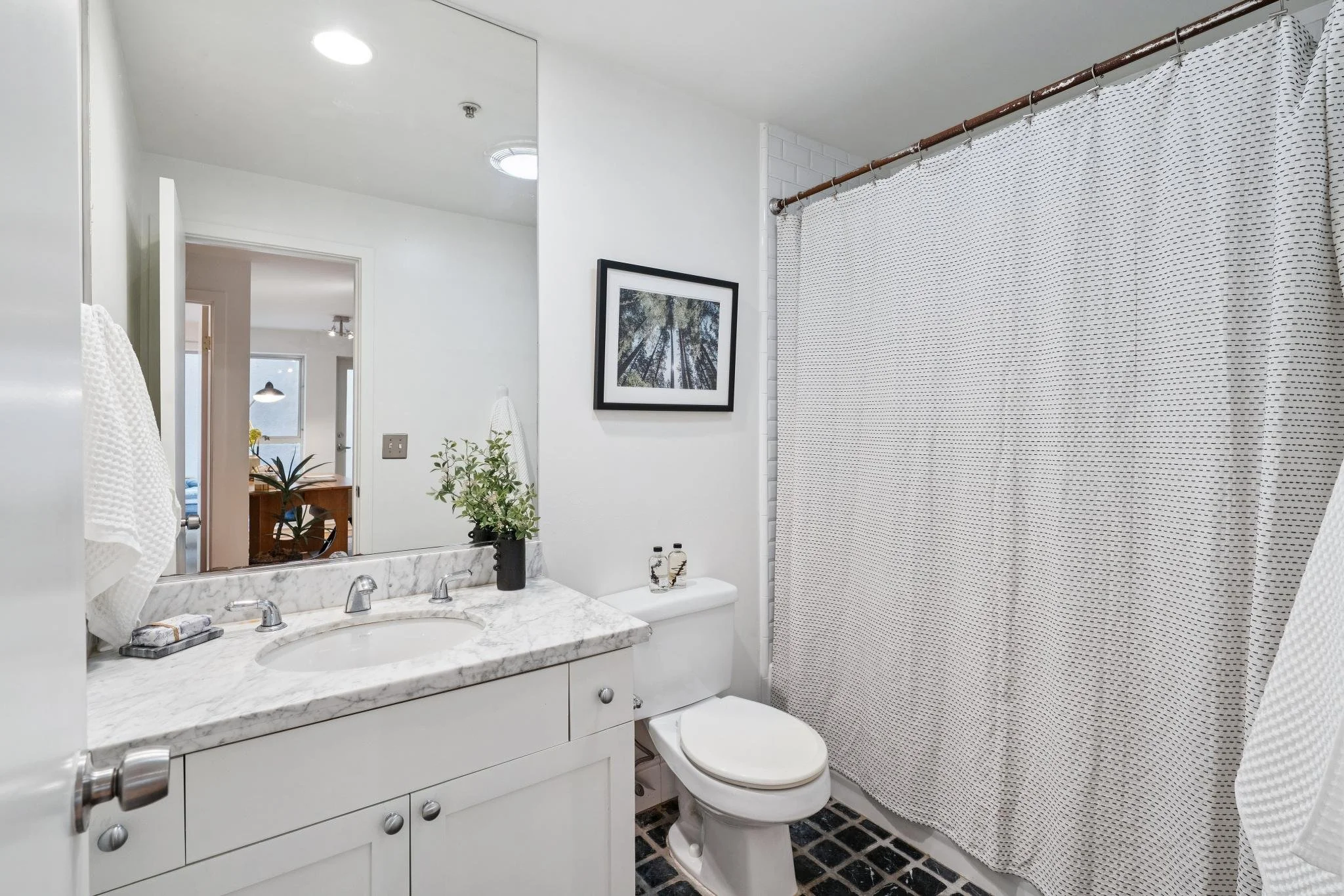 Bathroom with white vanity, marble countertop, and a toilet. There's a large mirror above the sink, a framed picture on the wall, and a shower area with a white curtain. The floor is tiled in black and white.