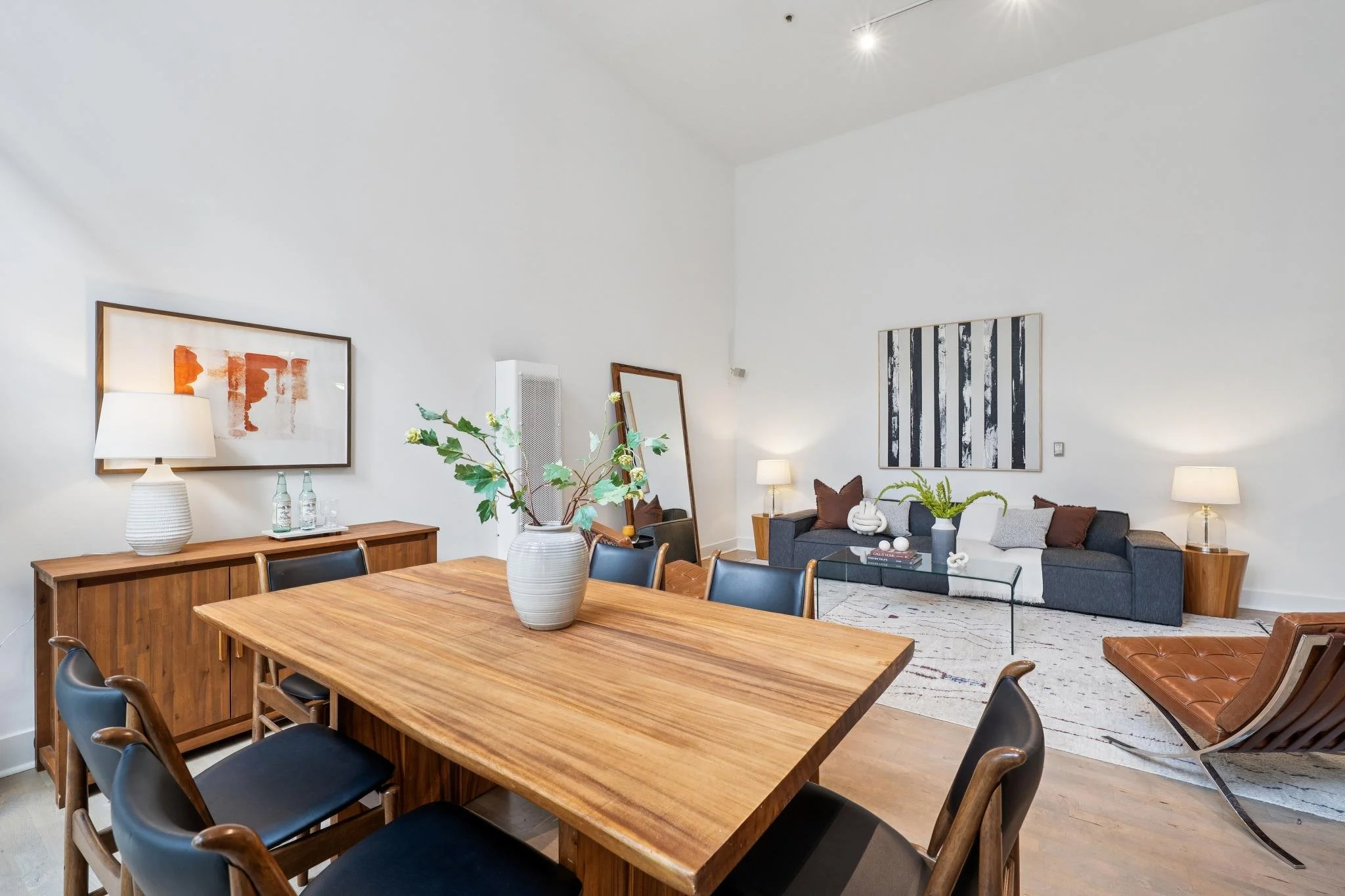 A modern dining and living room area with minimalist decor, wooden furniture, a gray sofa, black chairs, and artwork on white walls.