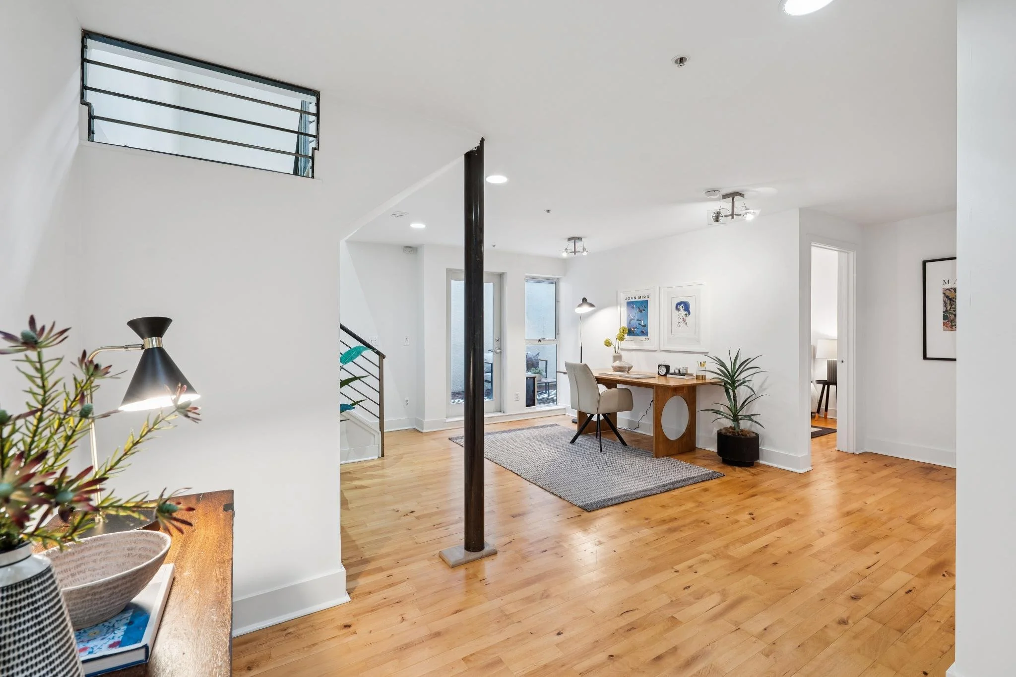 Open-plan living space with white walls, hardwood floors, a desk with a beige chair, potted plants, framed artwork, a sliding glass door, and a small enclosed balcony.