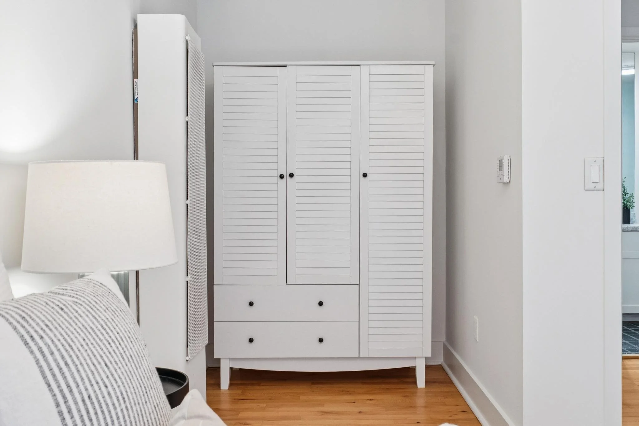 White wardrobe with louvered doors and drawers, next to a wall with thermostat and light switch, in a room with hardwood floor.