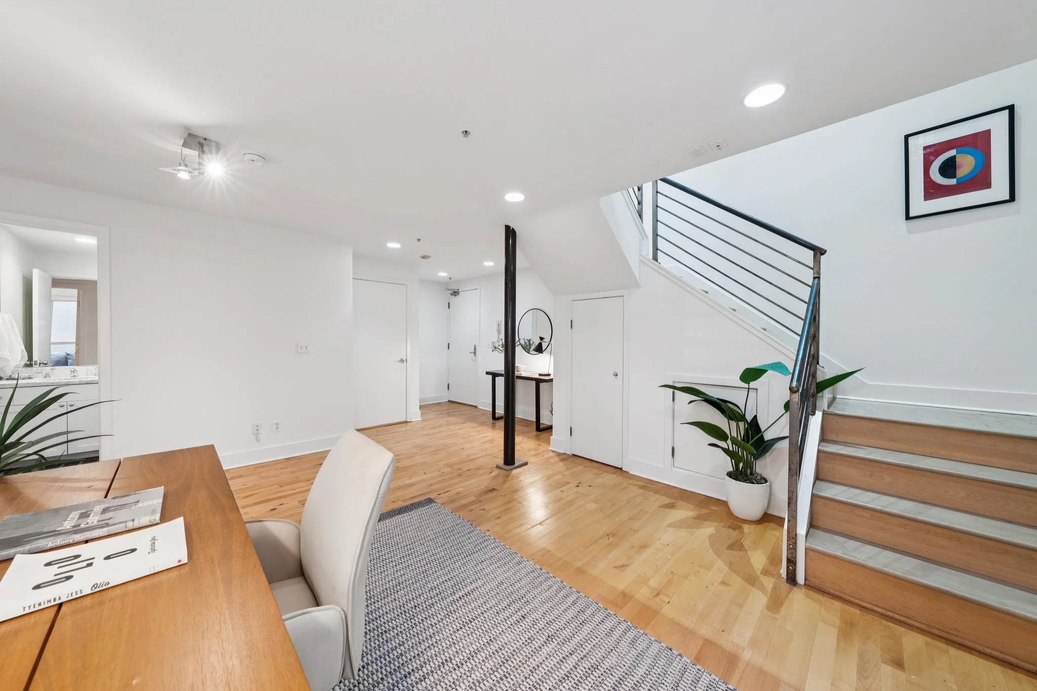 Living space with wooden floors, white walls, a staircase with metal railing, a potted plant, a painting on the wall, and a dining table with chairs.