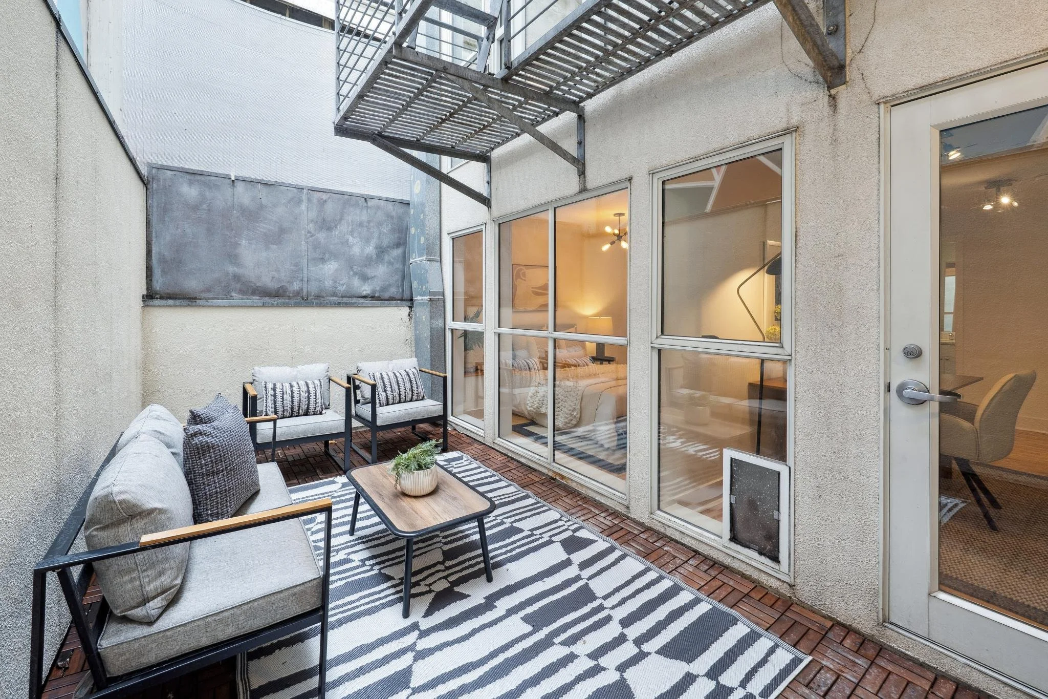 Outdoor patio with cushioned seating, striped rug, and small wooden table, enclosed by walls and large glass doors.