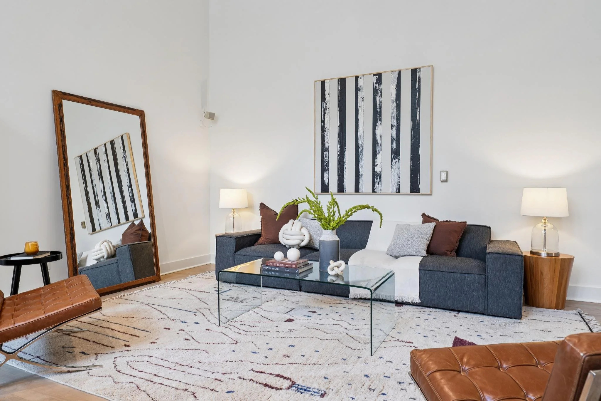 Modern living room with two dark gray sofas, a glass coffee table, a large mirror leaning against the wall, a large black and white striped artwork, a potted green plant, two table lamps with white shades, a white throw blanket, and decorative pillow