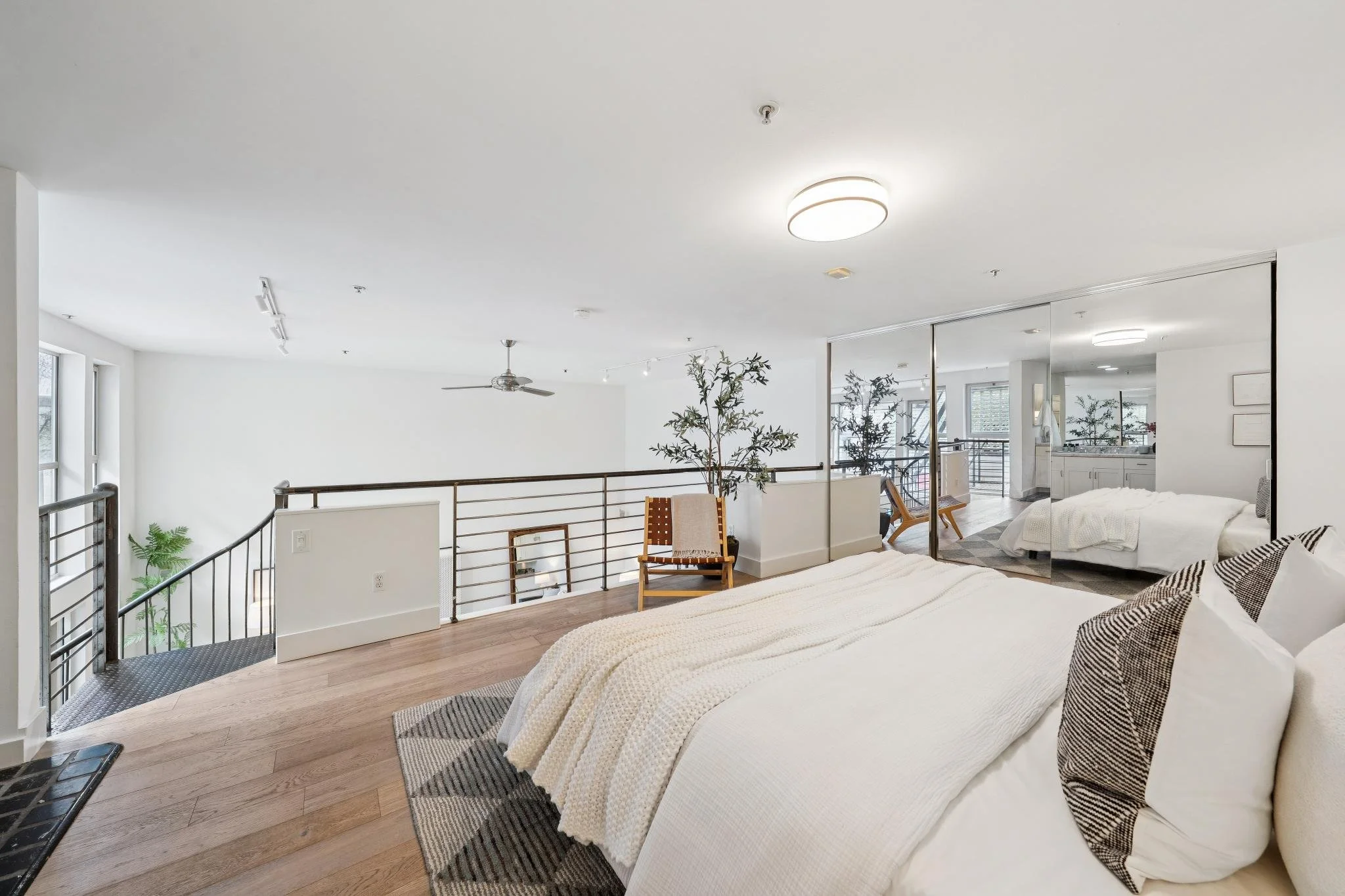 Bedroom with a bed, pillows, a blanket, mirrored closet doors, chairs, plants, and large windows allowing natural light.