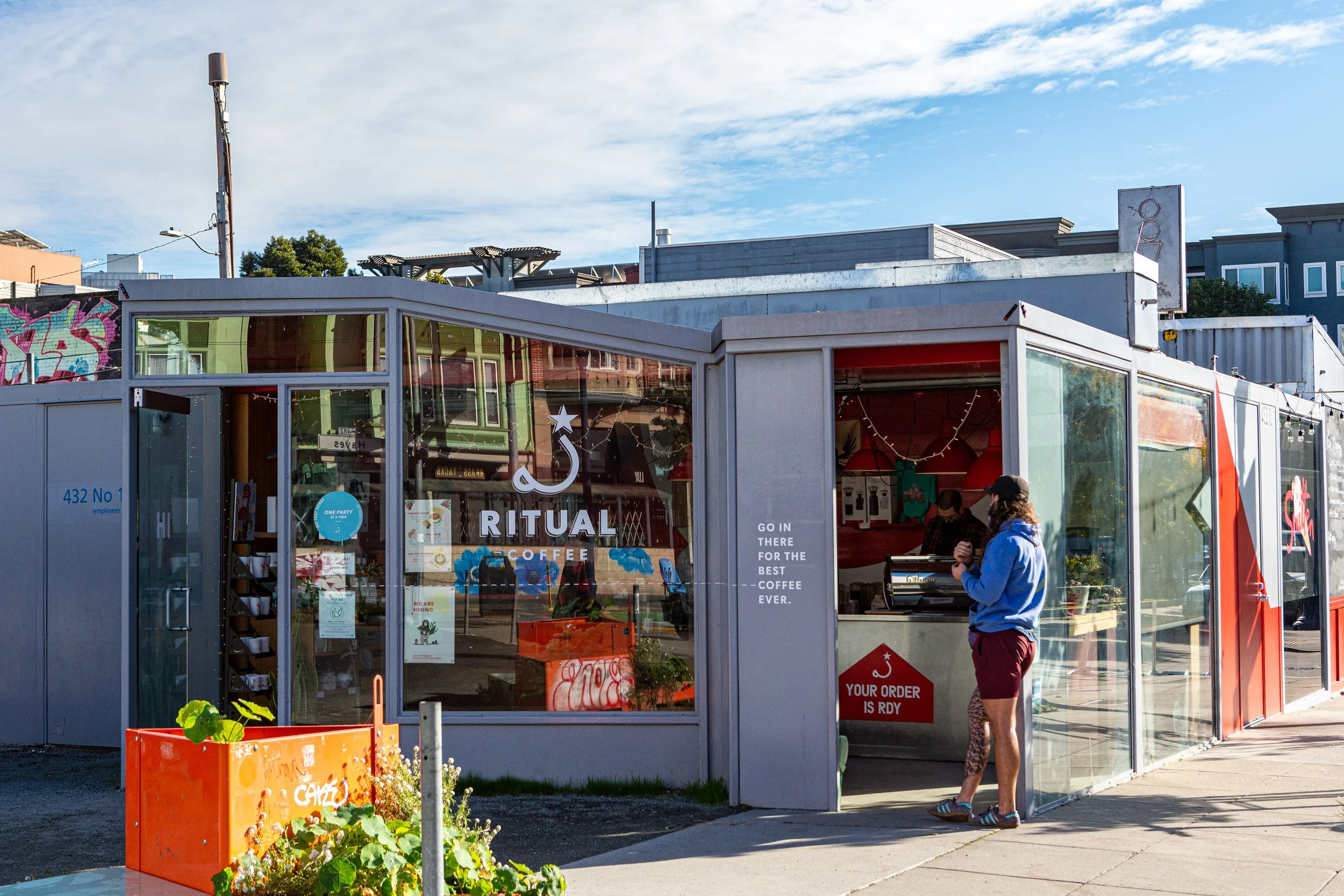 A small coffee stand named Ritual Coffee with large glass windows, a person in a blue hoodie and shorts ordering, and outdoor plants with an orange planter in front.