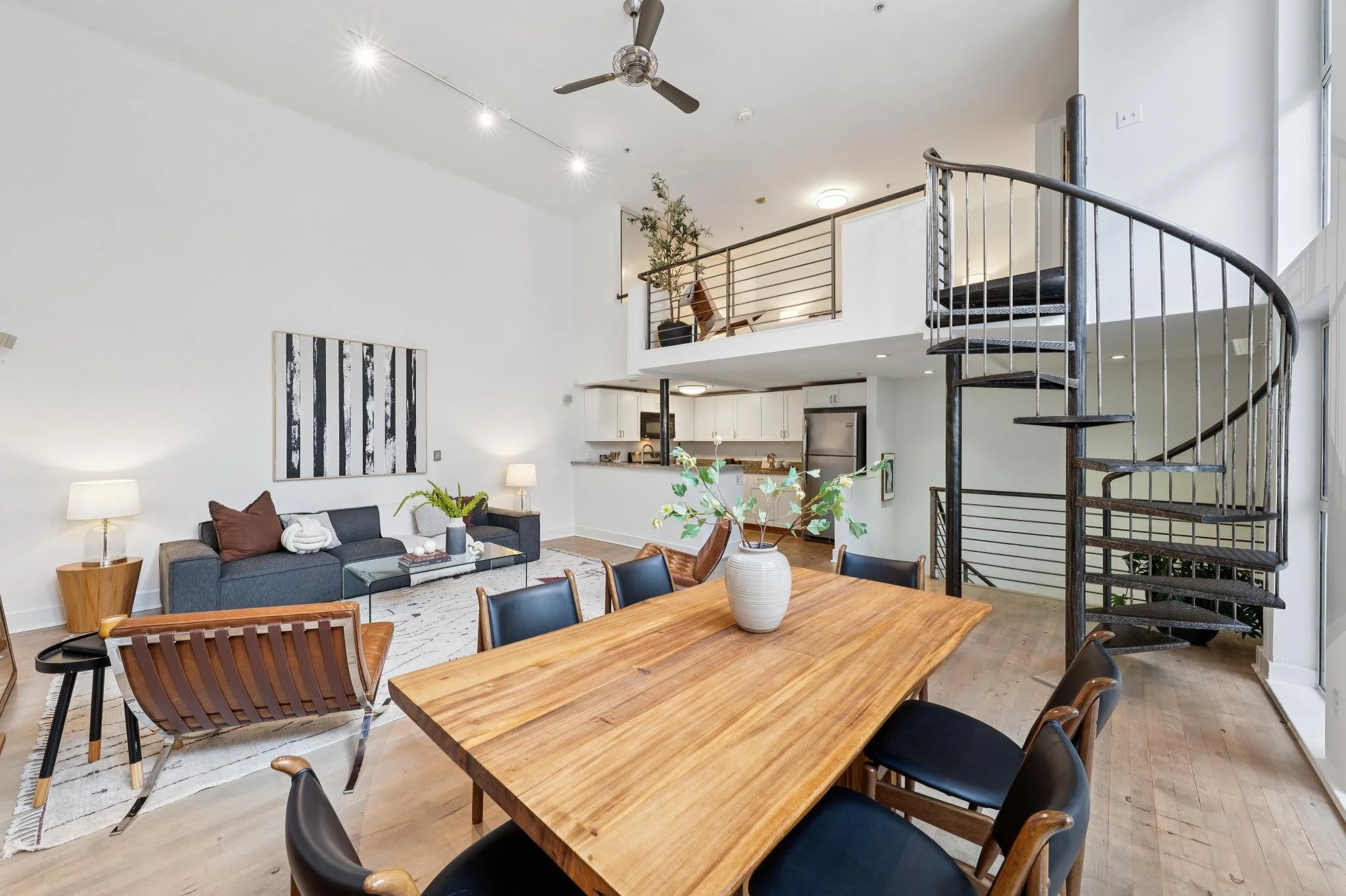 Open-concept living and dining area with a spiral staircase, kitchen, and modern furnishings including a wooden dining table, black chairs, and a blue sofa.