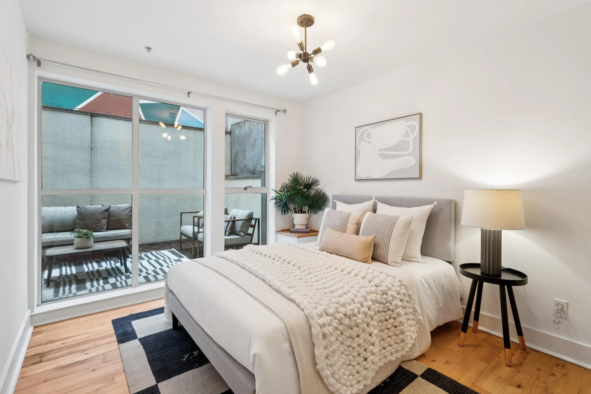 Modern bedroom with a large bed dressed in white, gray headboard, decorative pillows, a chunky knit throw blanket, a bedside lamp, a framed abstract art piece, and sliding glass doors leading to a small balcony with outdoor seating.