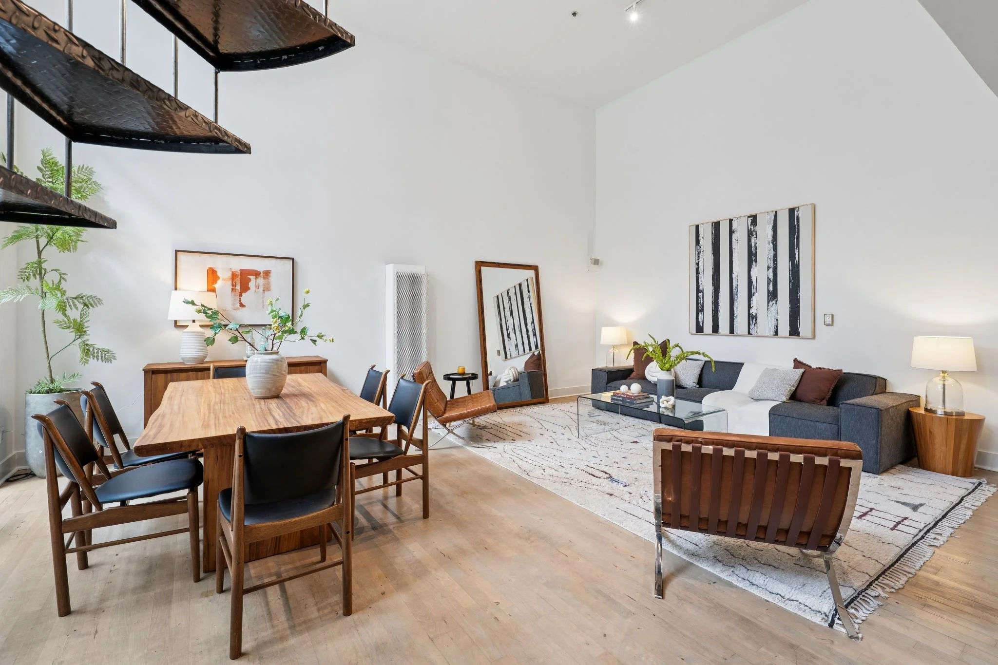 Contemporary living room with a wooden dining table and black chairs, a large gray sectional sofa, a tall mirror, abstract artwork, and various decorative elements on a patterned rug.