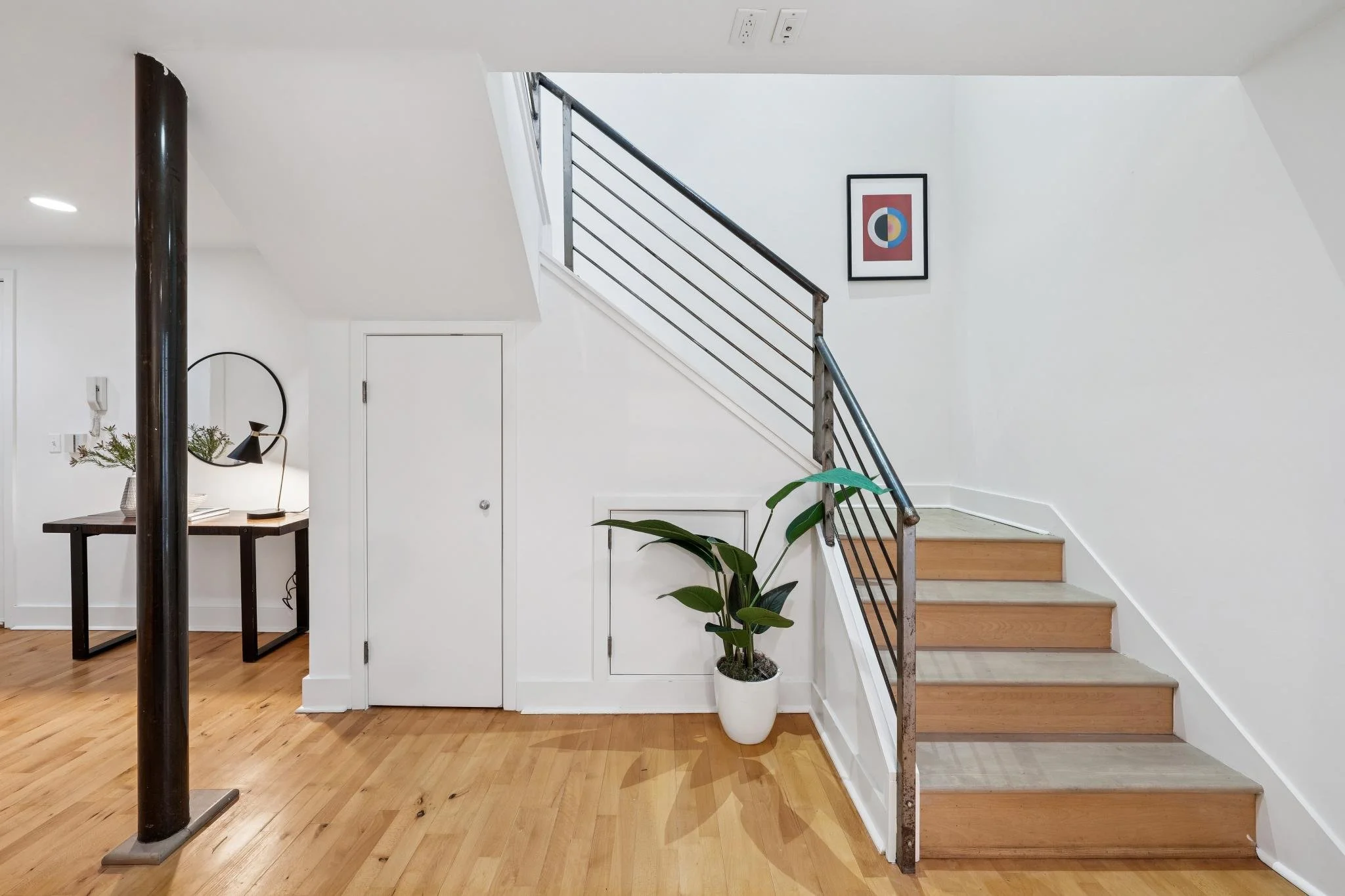 A modern interior with a staircase, a potted plant, a small desk with a mirror, and framed abstract art on the wall.
