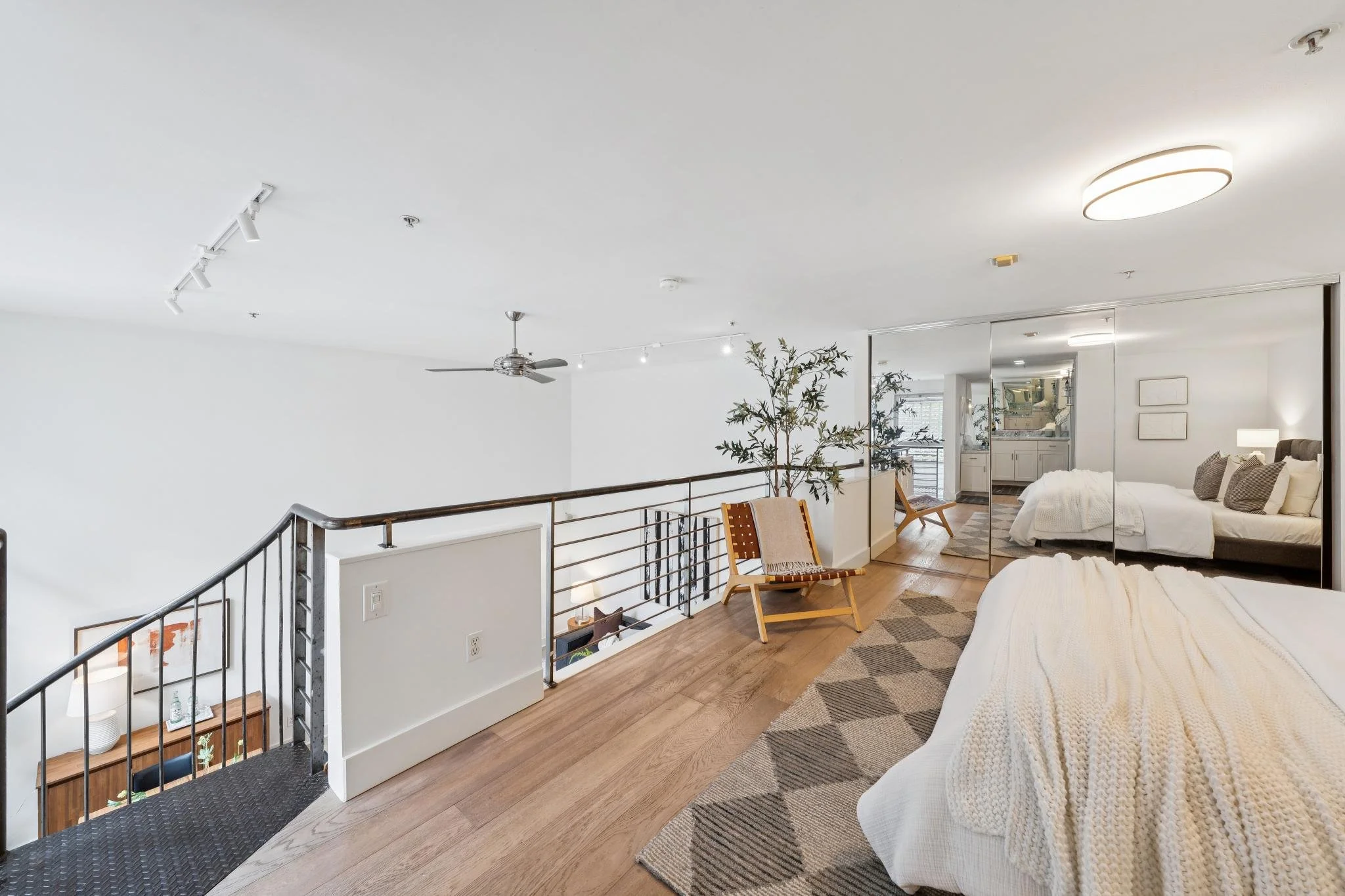A modern loft-style bedroom with mirrored closet doors, a bed dressed in white linens, sitting on a geometric rug, a comfortable chair, a large mirror, and a small dining area with a kitchenette in the background.