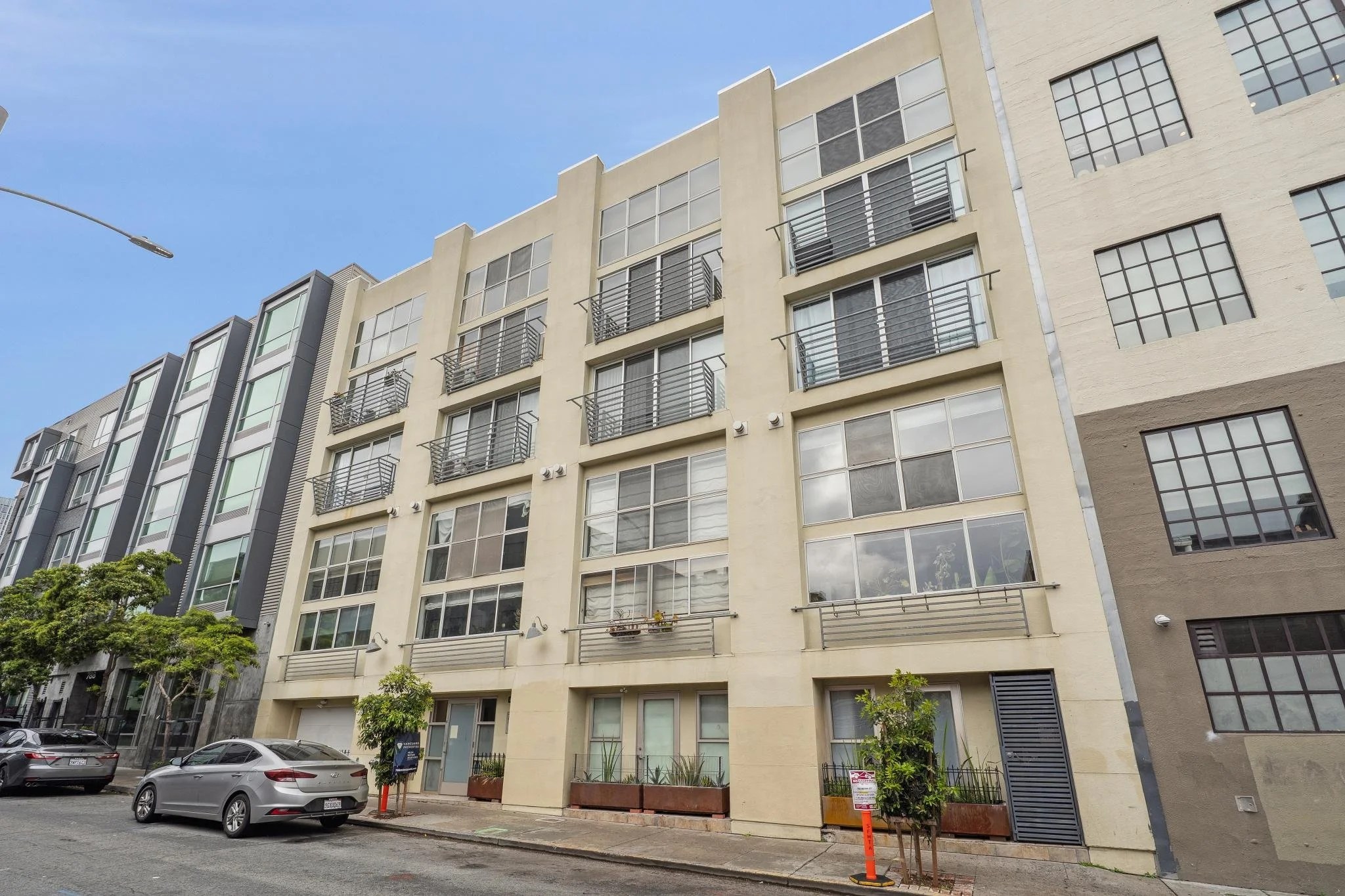 Multistory residential building with large windows and small balconies, parked cars on the street, a clear blue sky overhead, and trees lining the sidewalk.