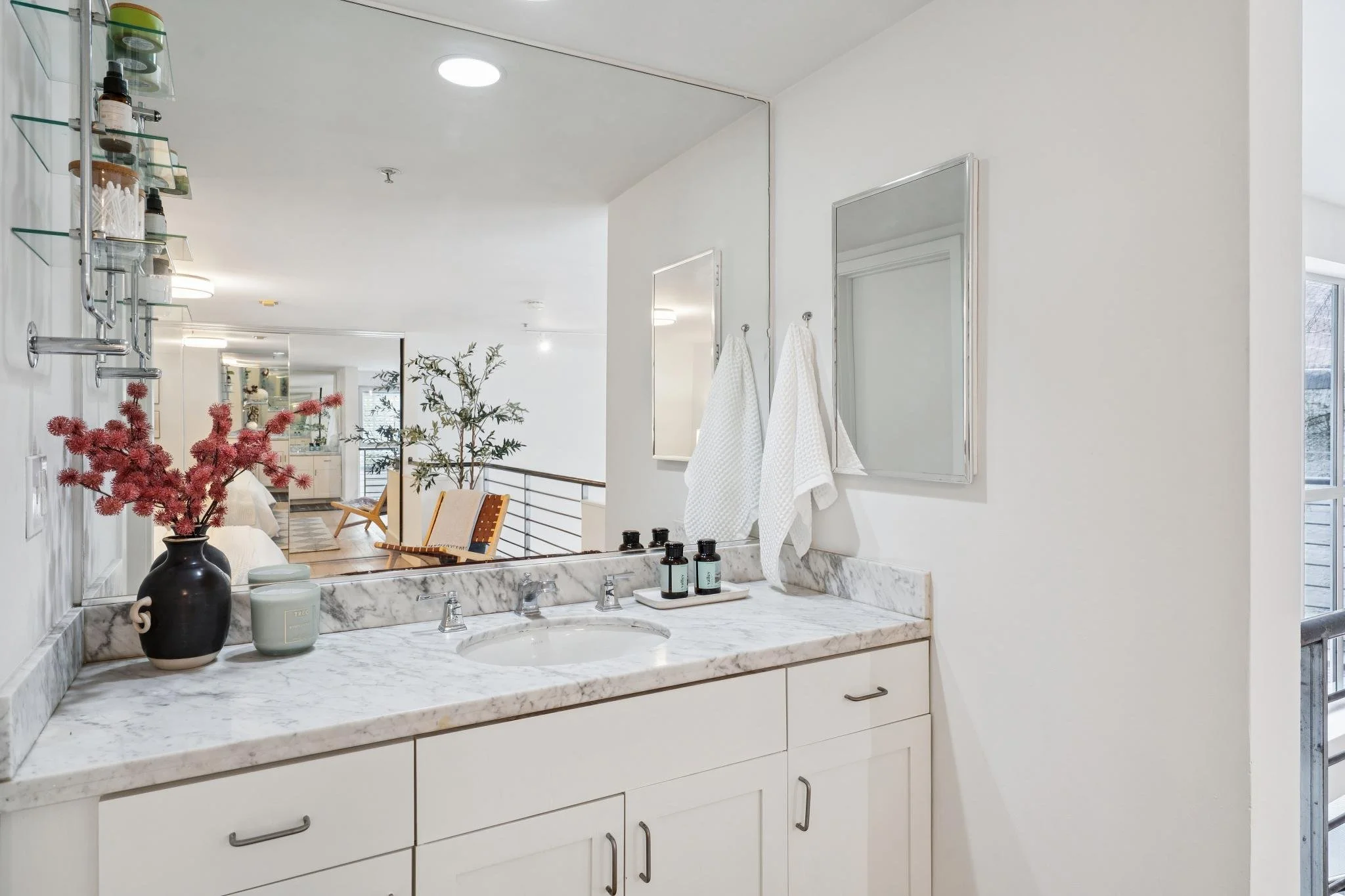 Bathroom vanity with white cabinets, marble countertop, a sink, and a large mirror. Decor includes a vase with red and pink flowers, a small container, and three bottles of toiletries. Towels hang on hooks beside a small mirror, and the background sh