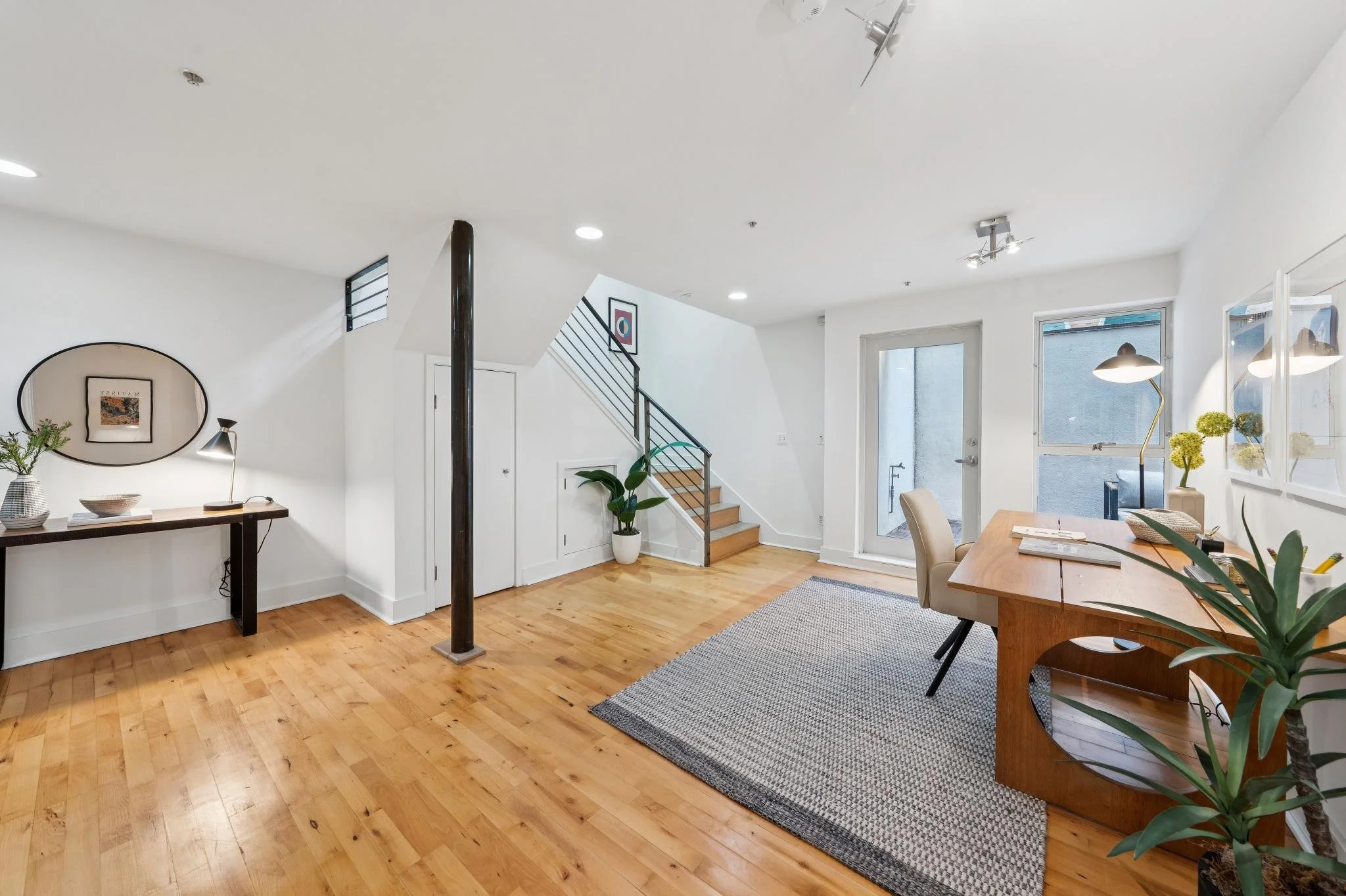 Interior of a modern room with light wooden flooring, white walls, a wooden staircase with black railing, a glass door, and a large window. There is a wooden desk with potted plants and books, a beige chair, a gray rug, and decorative wall art and li