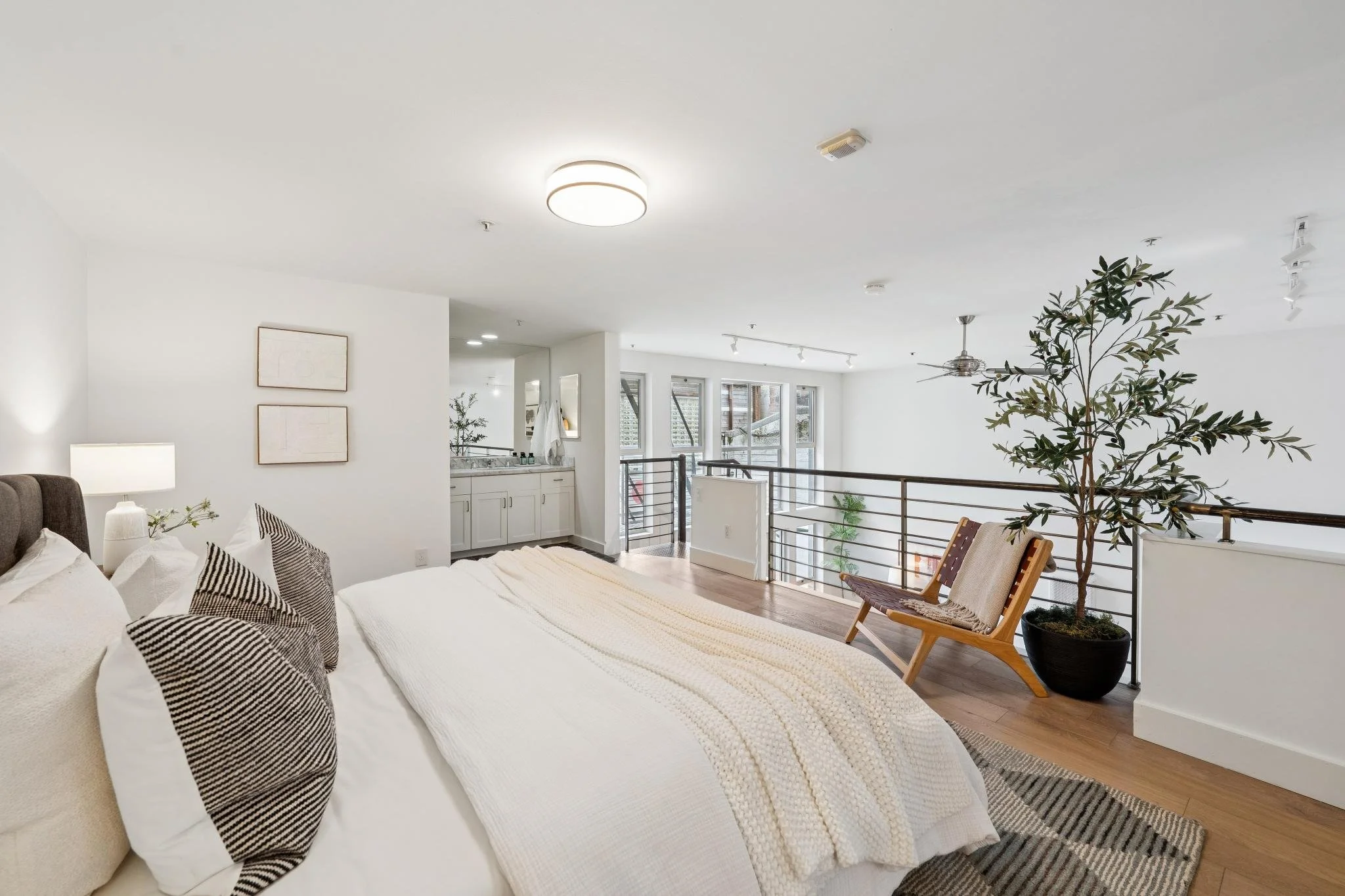 A modern bedroom with a large bed, pillows, and a throw blanket, a standing lamp, a chair with a blanket, and a large potted plant near a railing overlooking an open space.
