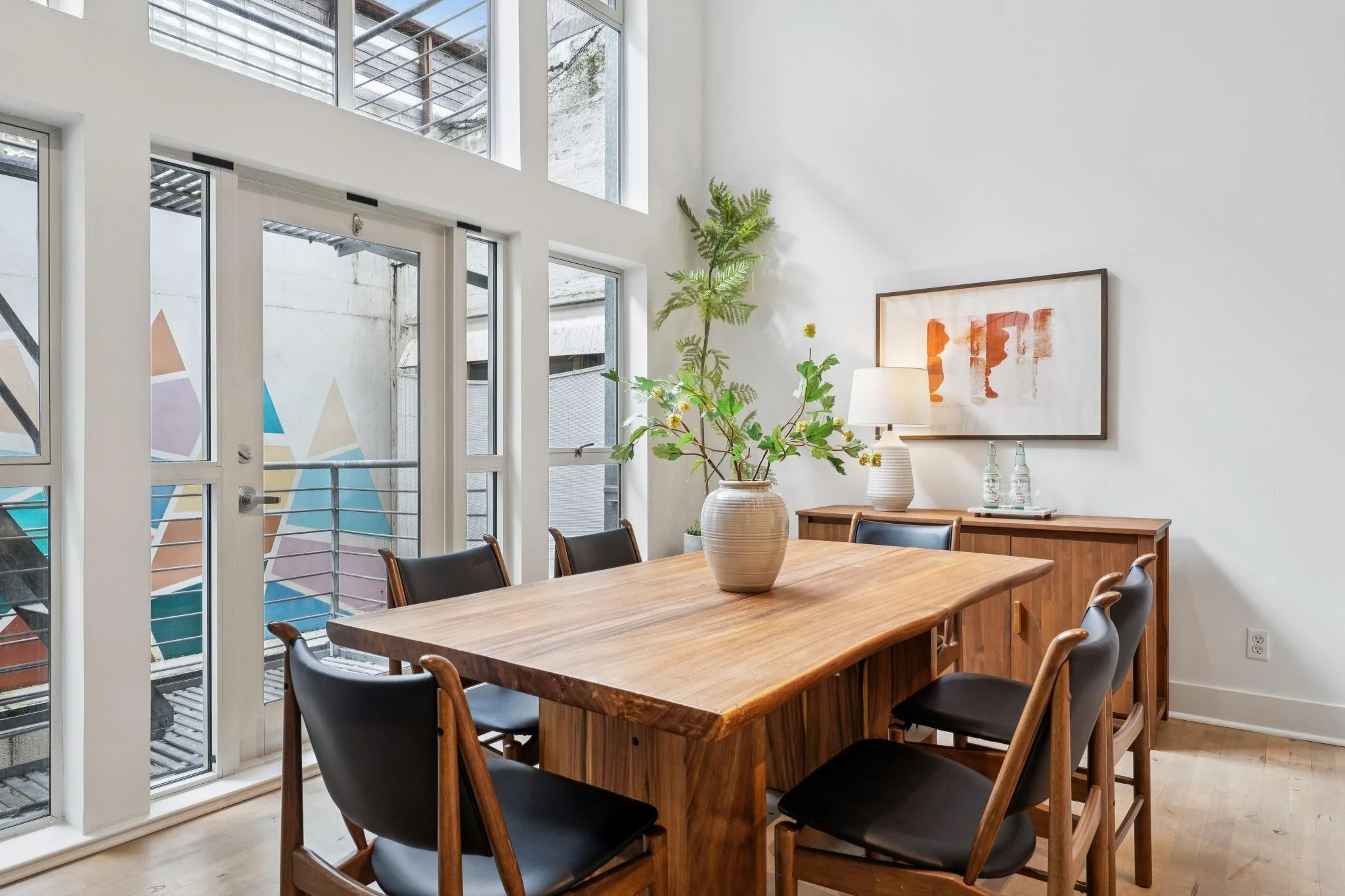 Modern dining room with a wooden table, black chairs, a vase with green foliage, artwork, and a sideboard with bottles and a lamp, next to large windows with a small balcony outside.