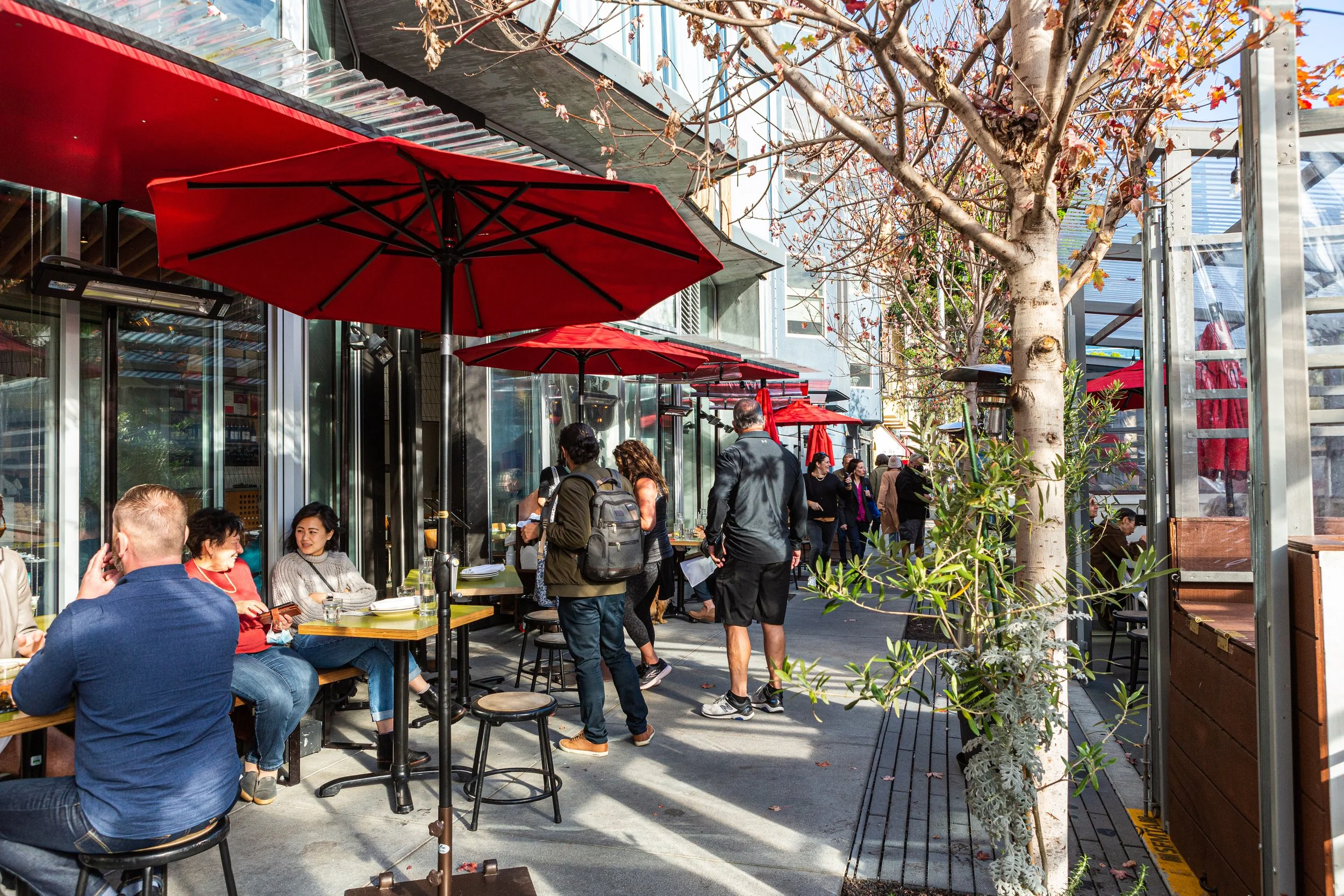 People dining outdoors at a cafe with red umbrellas, trees with autumn leaves, and a sidewalk