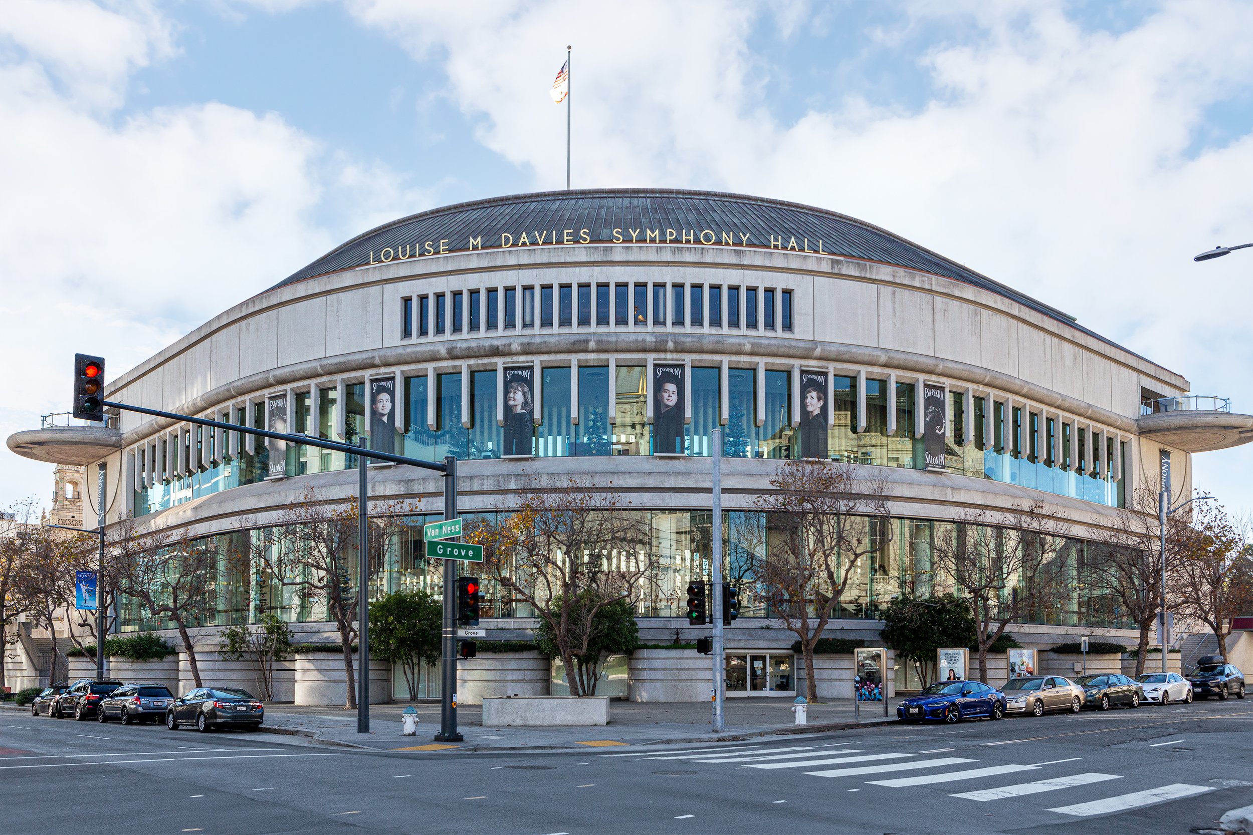 Louise M. Davies Symphony Hall in San Francisco, California, with visible street signs for Van Ness and Grove, traffic lights, parked cars, trees, and banners hanging on the building's windows.