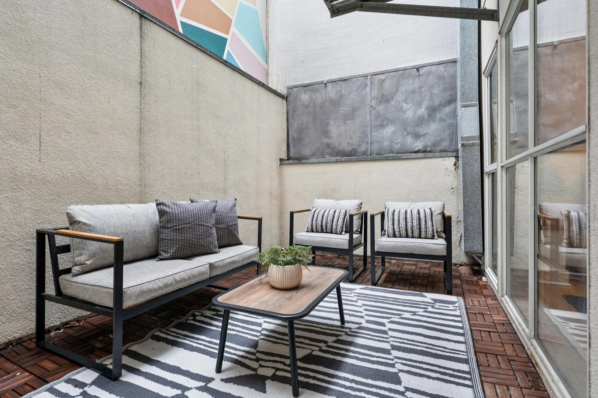 Outdoor patio with beige sofa and two armchairs with black frames, a wooden coffee table with a potted plant, black-and-white striped rug, and sliding glass doors.