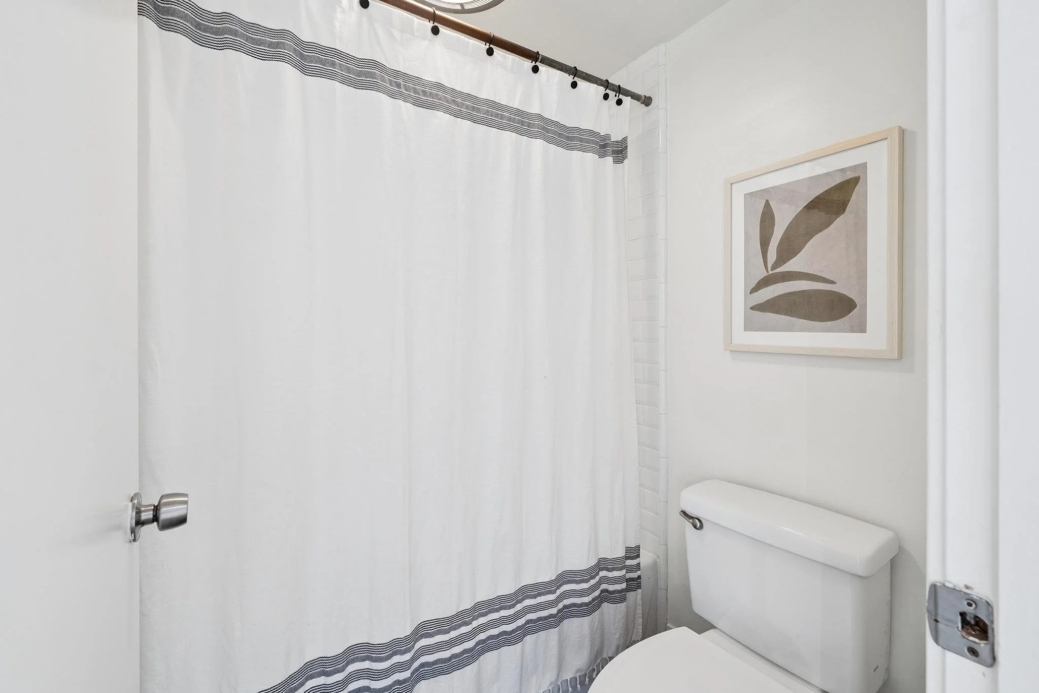 A small bathroom with a white toilet, a white shower curtain with gray stripes, and a framed abstract art piece on the wall.