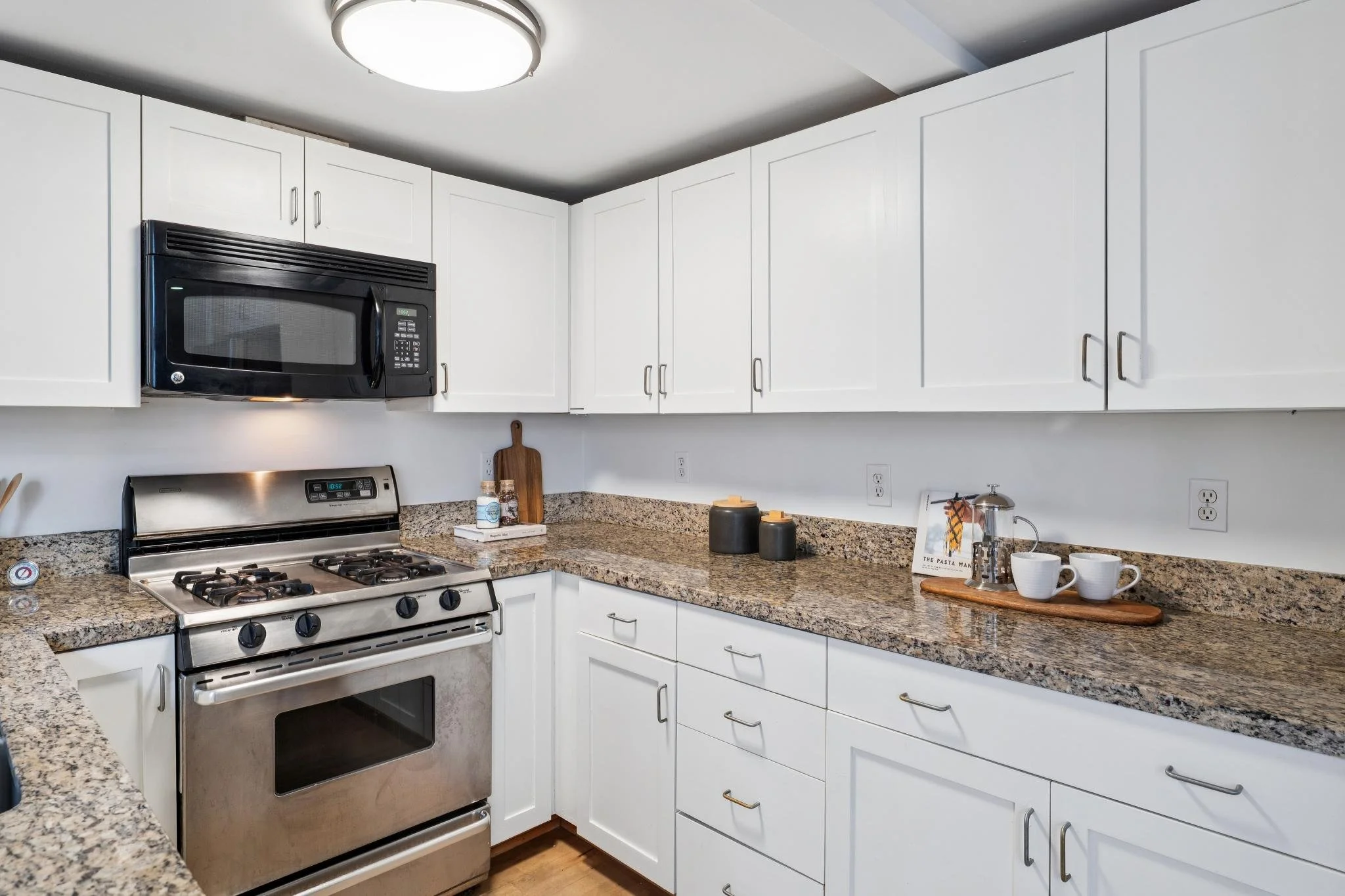 Kitchen with white cabinets, granite countertops, stainless steel gas stove, microwave above stove, and various kitchen items including cups, a cutting board, and decorative jars.