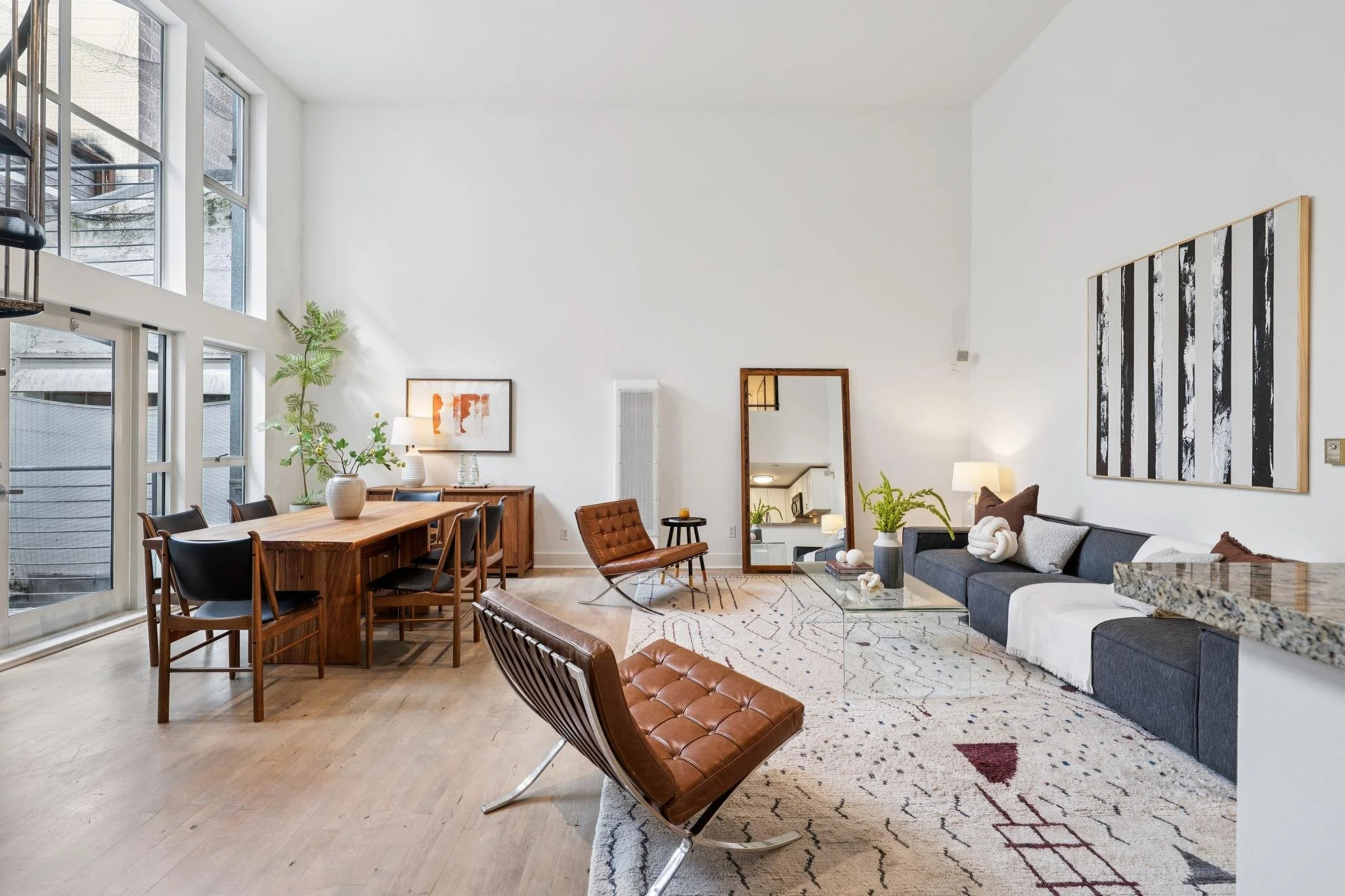 Modern living room and dining area with high ceilings, large windows, a gray sectional sofa, brown leather lounge chairs, a wooden dining table with black chairs, abstract artwork, and a mirror.