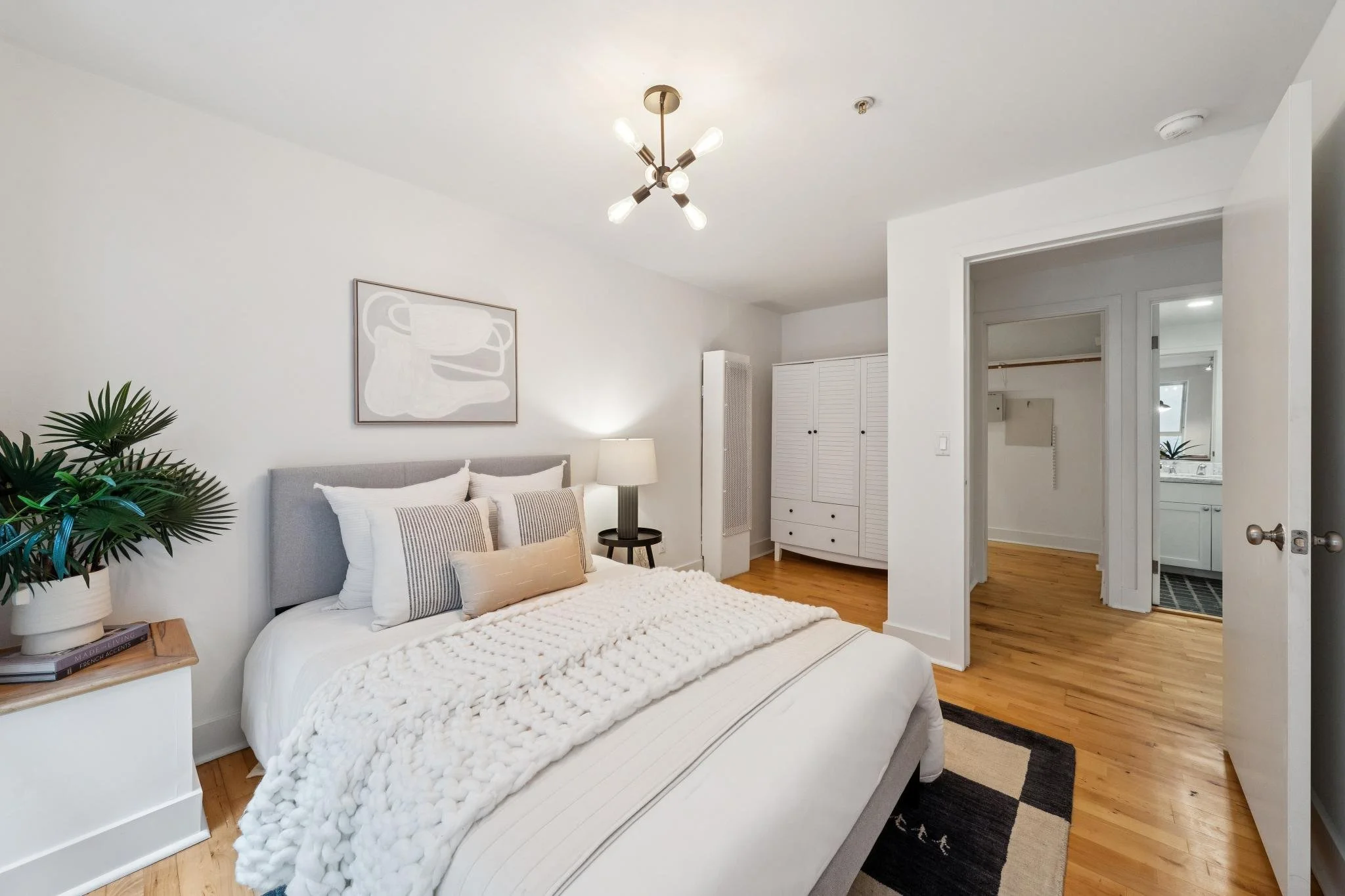 A bedroom with a white bed, gray headboard, beige and striped pillows, a white textured blanket. A potted plant on a white nightstand, a black table lamp, a framed abstract artwork of a bear, and a white wardrobe. Hardwood floors, open doorway to a b