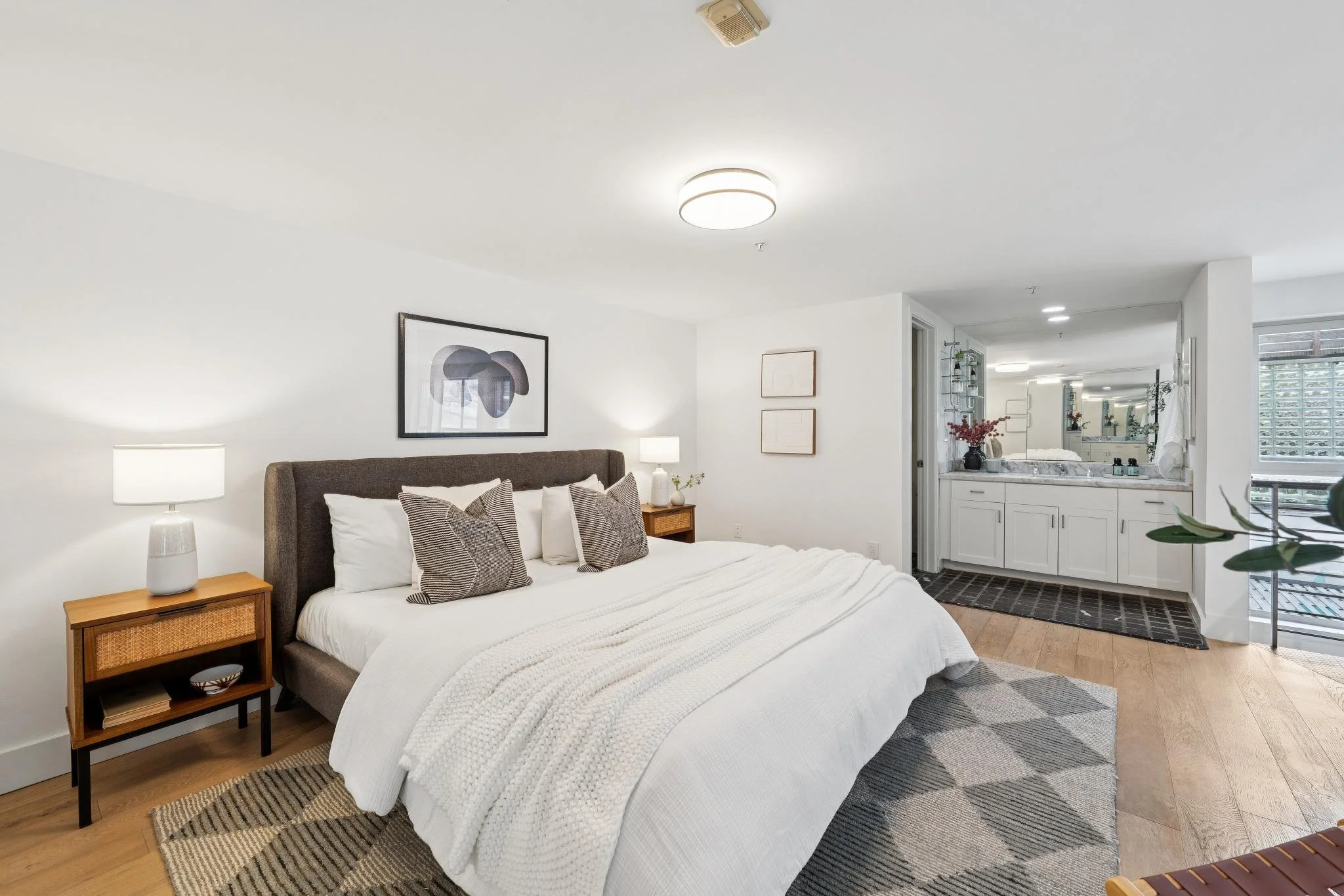 A bedroom with a large bed with white bedding and throw pillows, two bedside tables with lamps, wall art above the bed, a small rug at the foot of the bed, and a view into a bathroom with a vanity and mirror, with natural light coming through a windo
