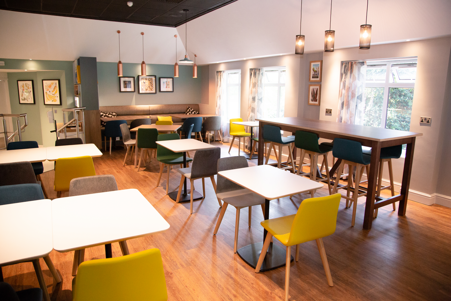 Interior of a modern, well-lit cafe with colorful chairs, white tables, pendant lighting, and framed artwork on the walls, with large windows allowing natural light.