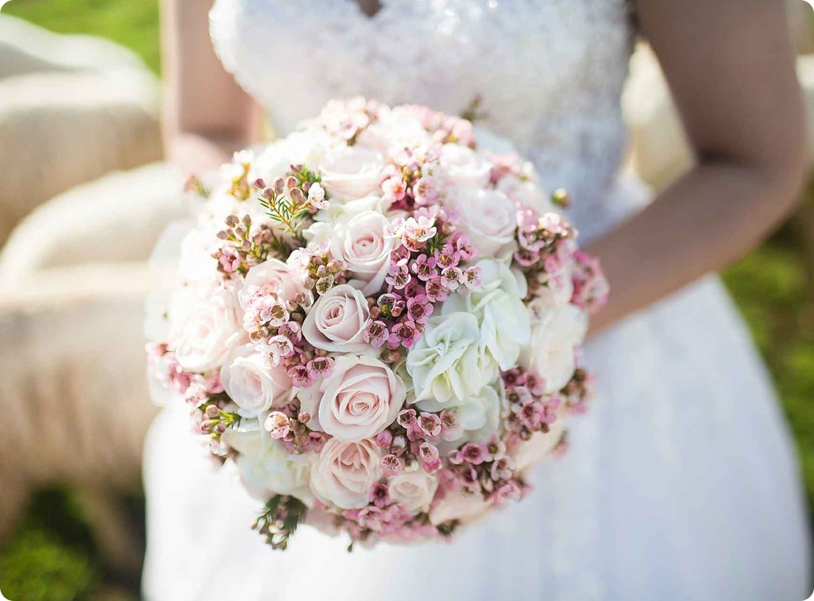 Close-up of a bride holding a bouquet of pink and white roses with small pink flowers, outdoors