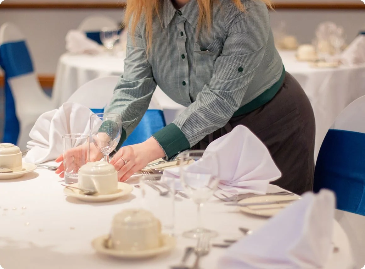 A woman is setting a dining table with white napkins, glassware, and dinnerware in a banquet hall or restaurant setting.