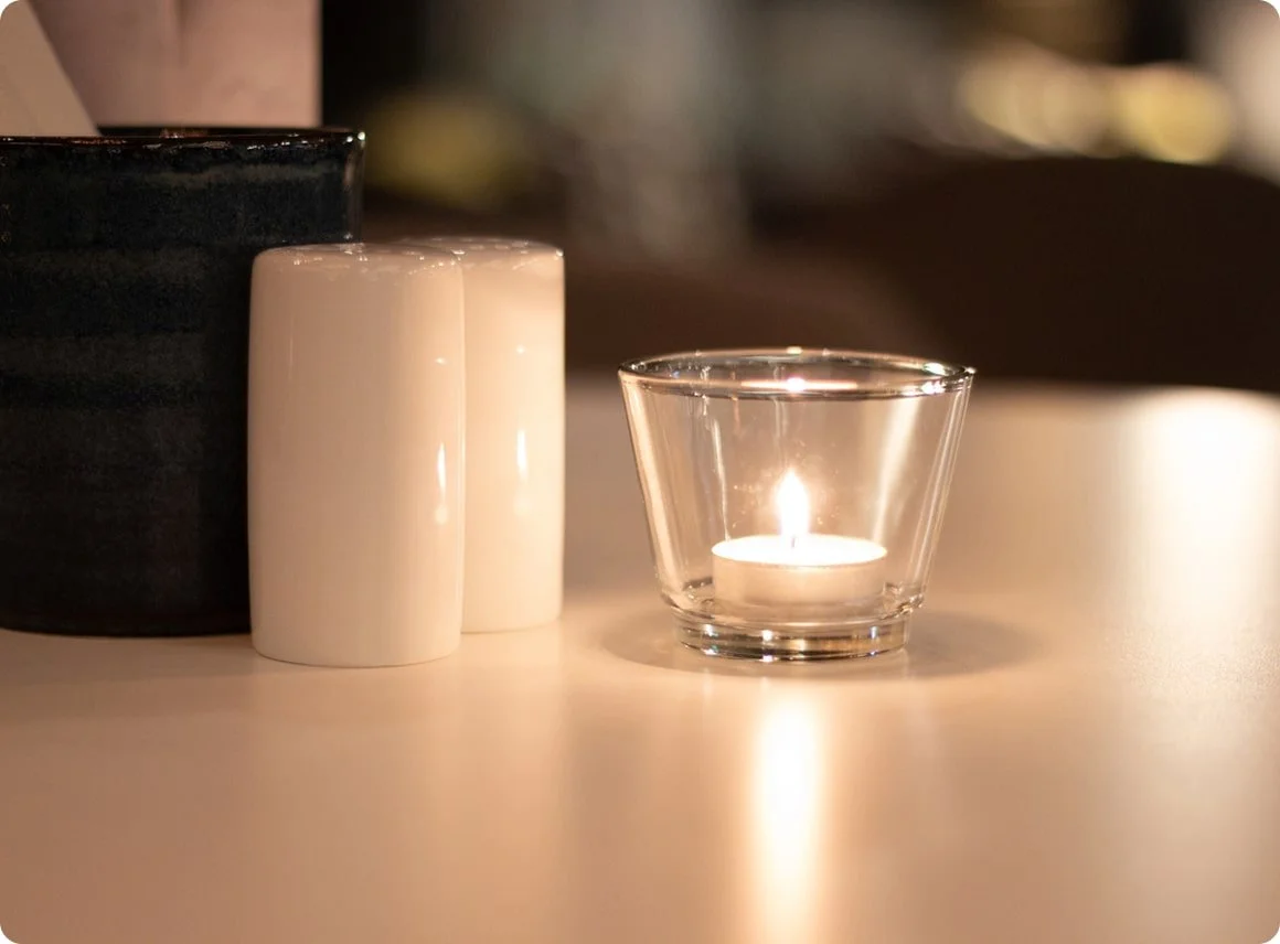 A lit tea light candle in a clear glass holder on a white surface, with two tall, white candlesticks and a black container in the background.