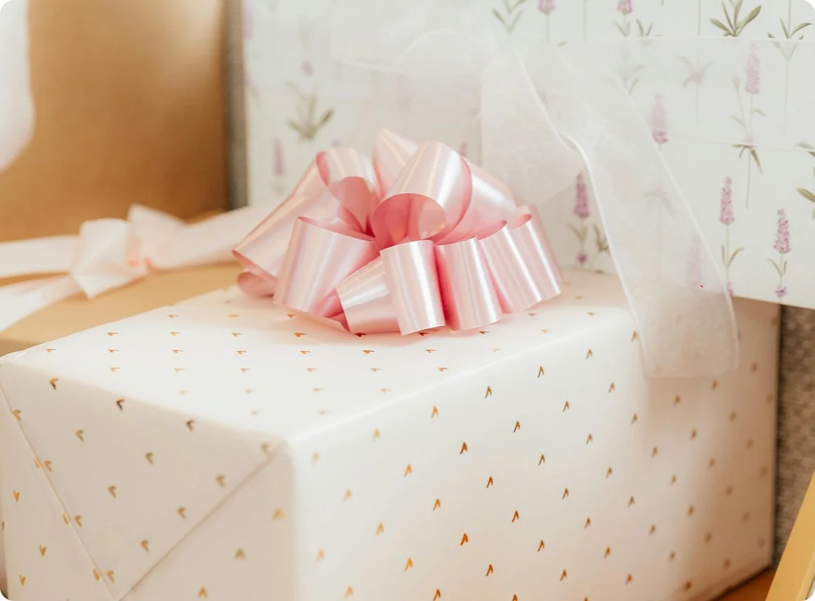 Decorative gift box wrapped in white paper with small gold arrow patterns, topped with a pink ribbon bow, surrounded by other wrapped gifts and floral-patterned wrapping paper.