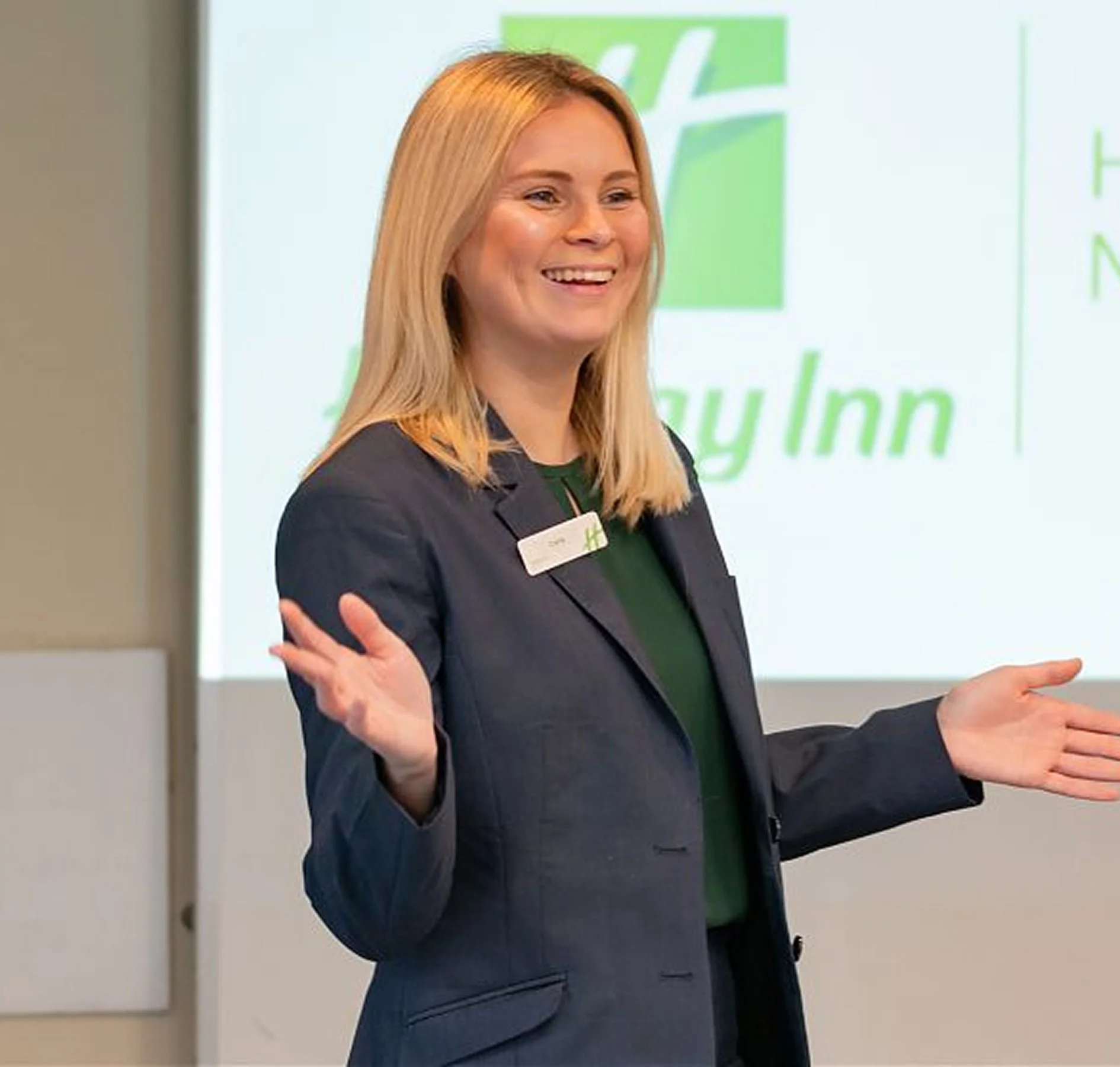 A woman with long blonde hair, wearing a dark blazer and green top, speaking in front of a screen with the word 'BayINN' visible, gesturing with her hands during a presentation.