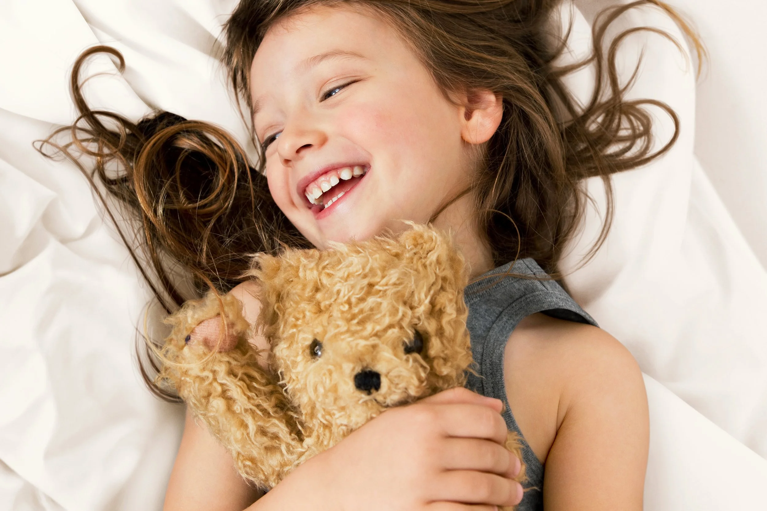 A young girl with long, curly brown hair smiling while lying on a white surface, holding a plush teddy bear.