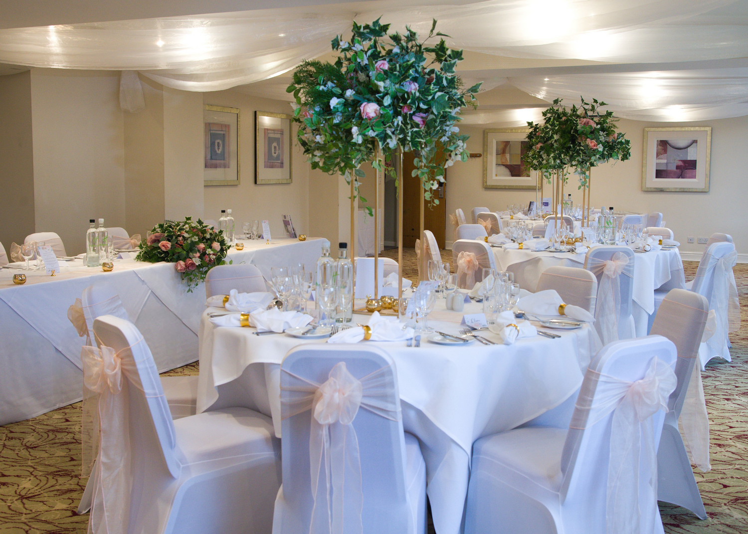 Elegant banquet hall decorated with round tables covered in white tablecloths, adorned with floral centerpieces, set with glassware, cutlery, and napkins. Chairs are dressed with white covers and peach-colored bows. The ceiling has draped fabric, and framed artwork decorates the walls.