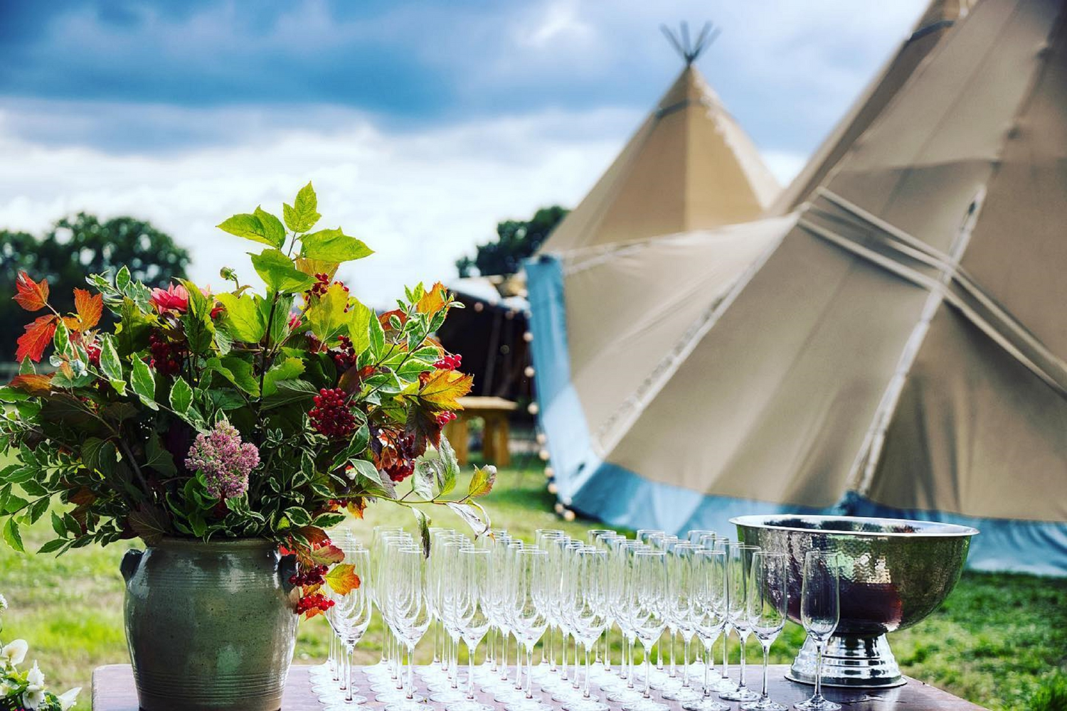 A table with a large floral arrangement, multiple champagne glasses, and a silver ice bucket with red wine, set outdoors with a large tipi tent and cloudy sky in the background.