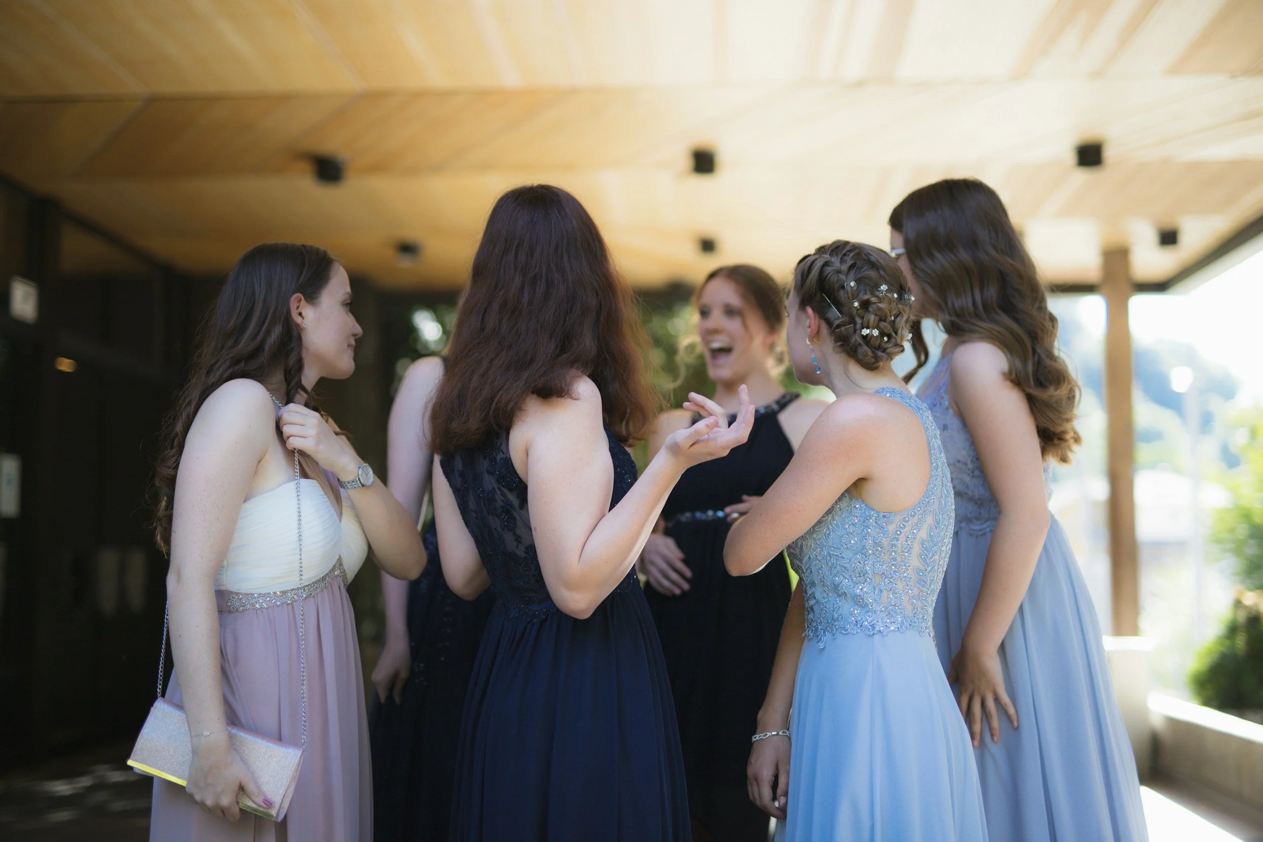A group of six women in formal dresses are talking and laughing outdoors under a wooden overhang.