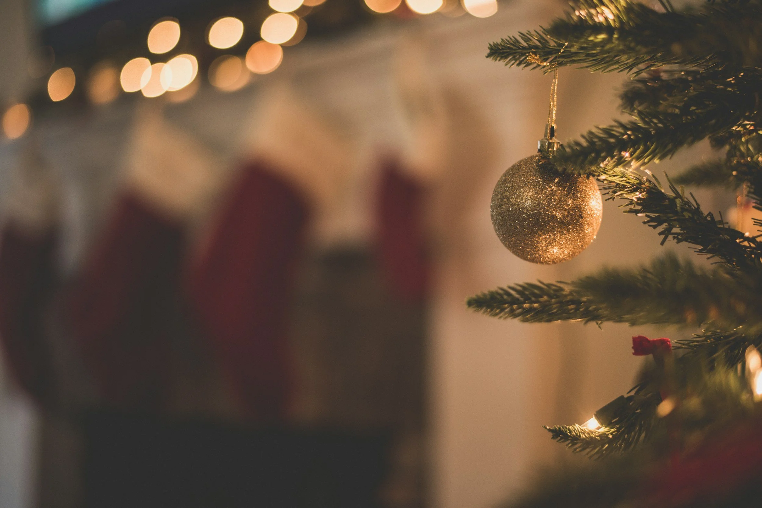 A close-up of a decorated Christmas tree branch with a gold glittery bauble ornament, with warm bokeh lights in the background.