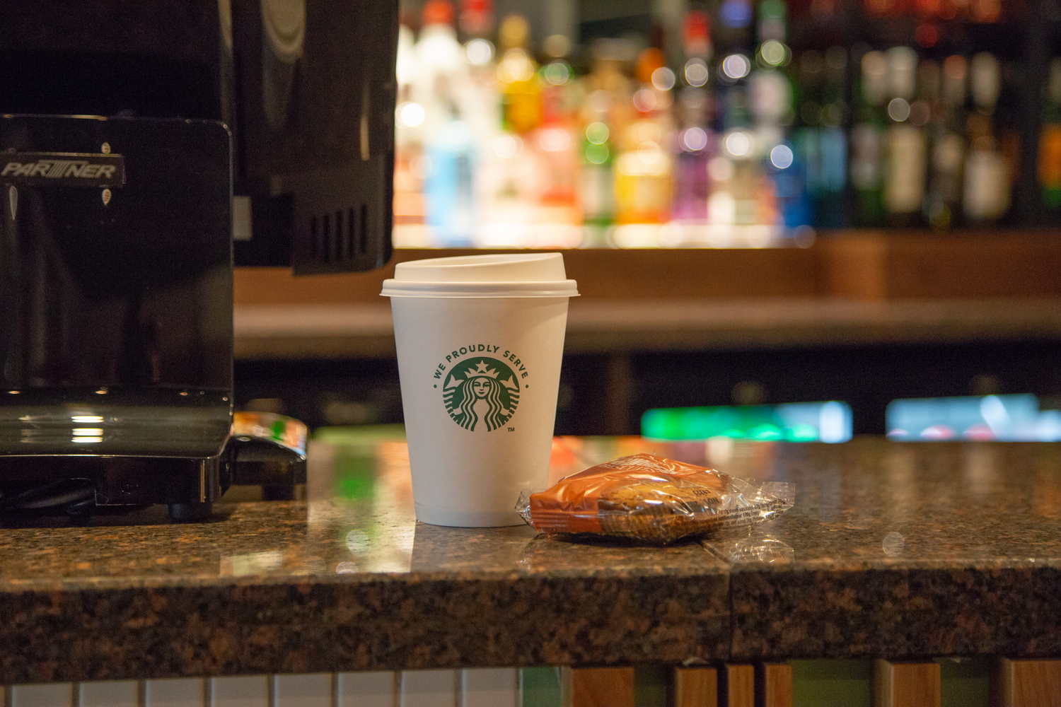 A Starbucks paper coffee cup with a white plastic lid and a wrapped pastry sit on a granite countertop inside a cafe, with a blurred background of colorful bottles on shelves.