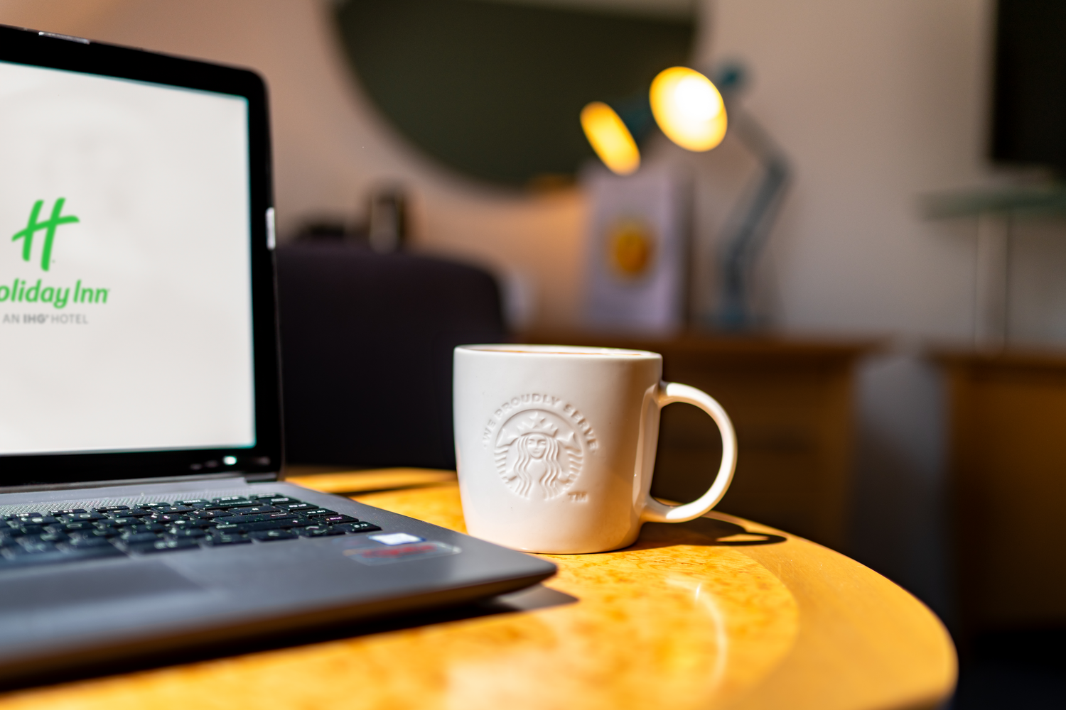 A white Starbucks coffee mug on a wooden table next to a laptop displaying a Holiday Inn logo on the screen. In the background, a desk lamp with yellow lights is visible.