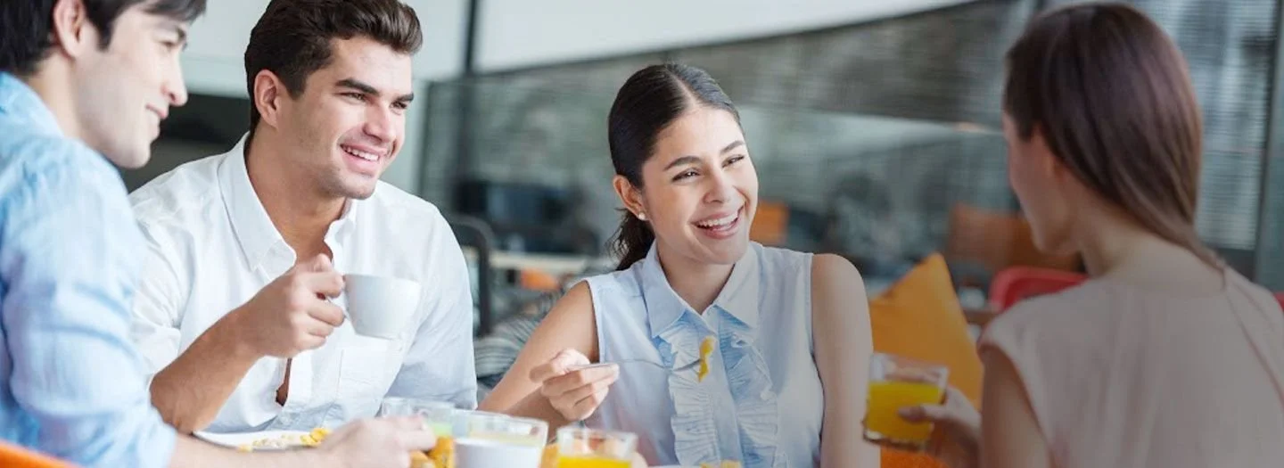Four young adults sitting at a table in a bright, modern cafe, engaging in conversation and enjoying beverages, with a woman smiling in the center of the group.