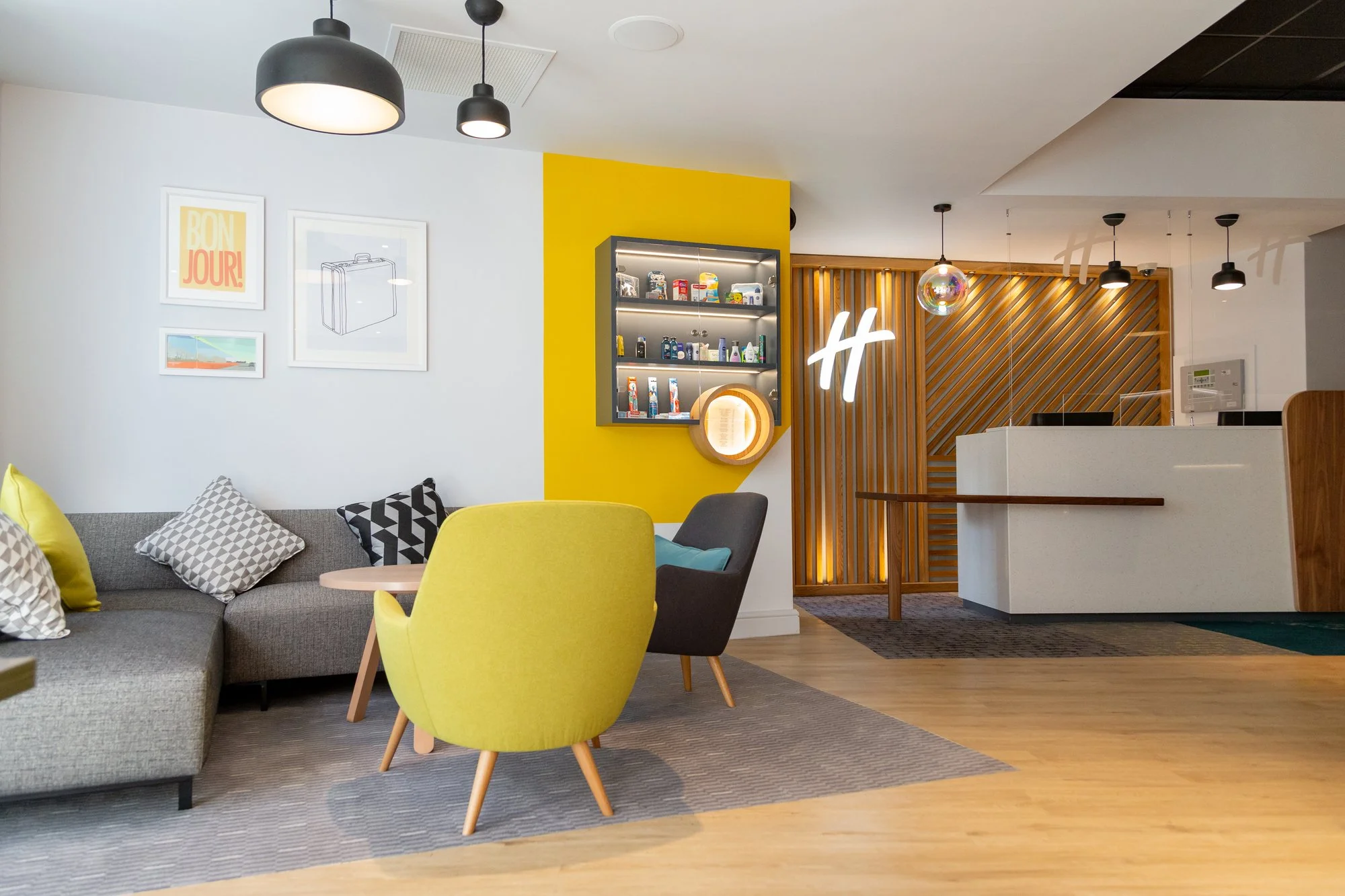 Interior of a modern hotel lobby with a gray sofa, yellow and gray chairs, framed art on white walls, a yellow accent wall with a display cabinet, and a white reception desk. The ceiling has black pendant lights and a decorative disco ball.