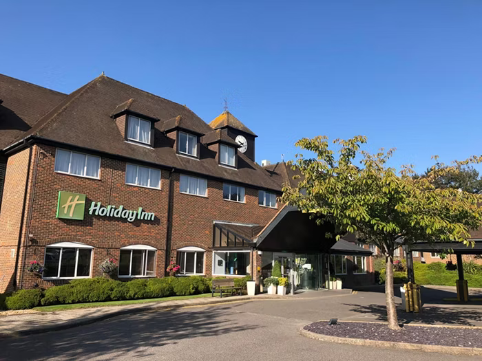 Exterior view of a Holiday Inn hotel with a brick facade, multiple windows, and a clock tower, surrounded by trees and a parking area on a sunny day.