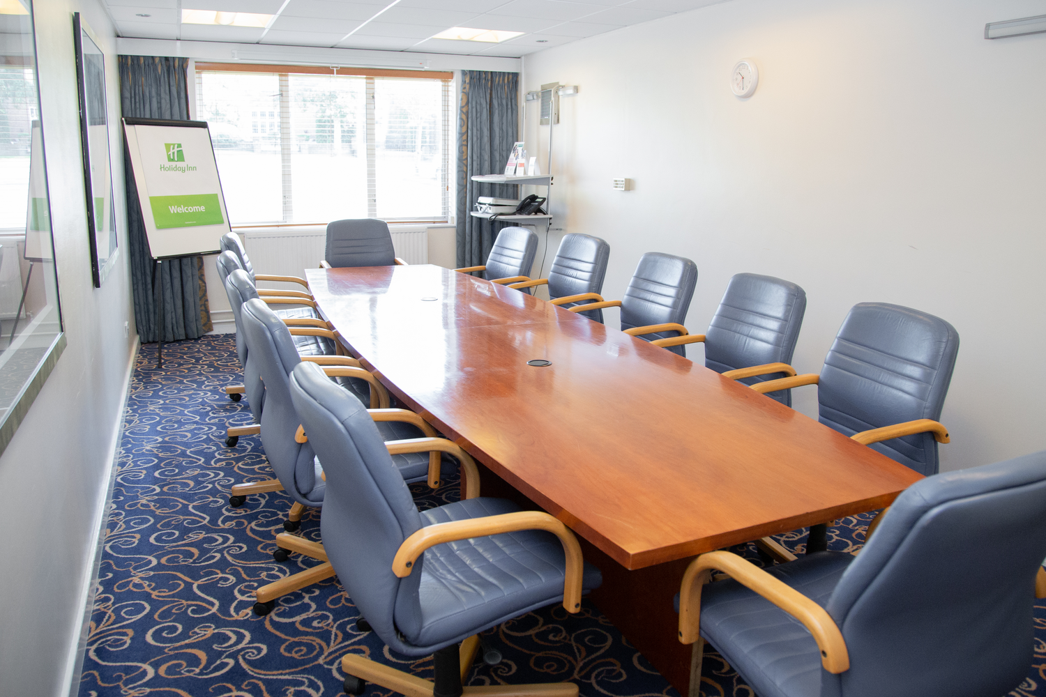 A conference room with a large wooden table surrounded by ten blue office chairs, and a window with curtains. There is a Holiday Inn welcome sign on a stand near the wall and a shelf with a phone, brochures, and some equipment in the background.